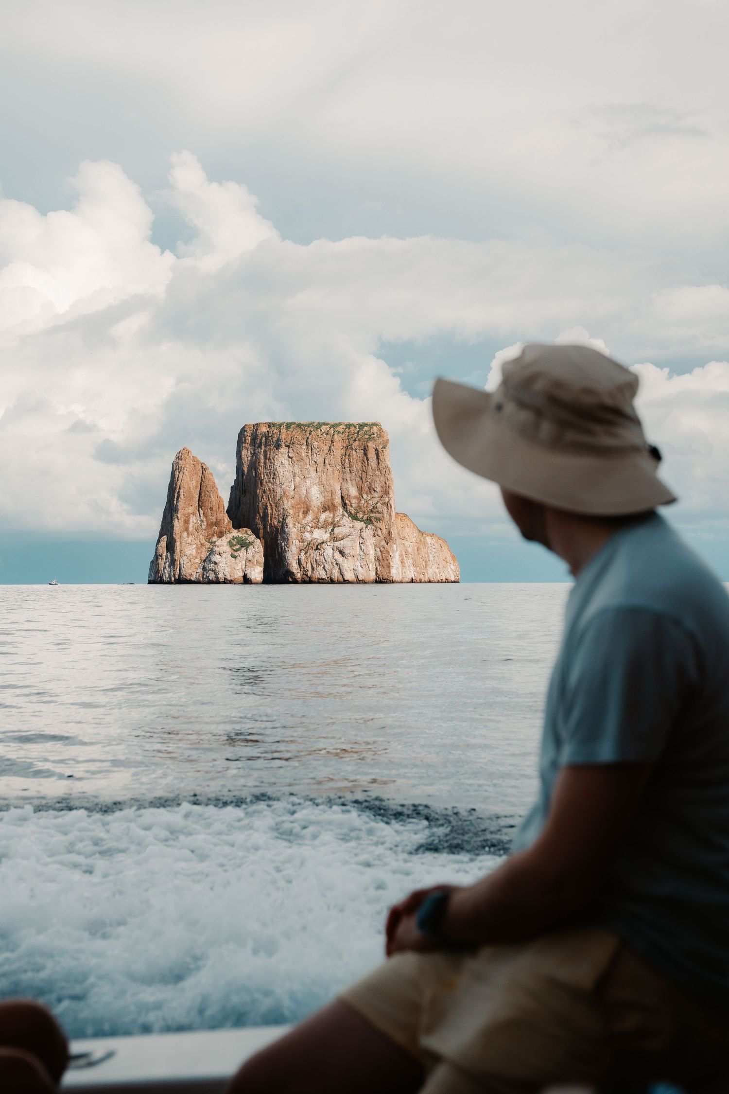 Man in sun hat sits on boat gazing at dramatic rocky sea stack rising from calm ocean waters under cloudy sky.