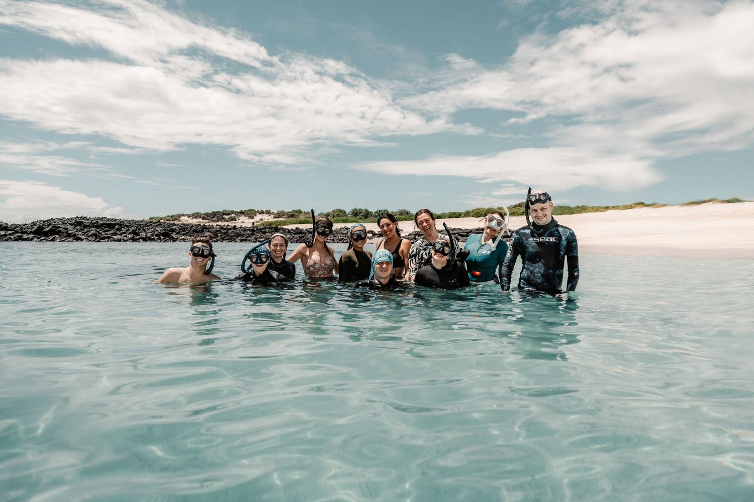 Group of snorkelers posing together in crystal clear shallow waters near a sandy beach under a partly cloudy sky.