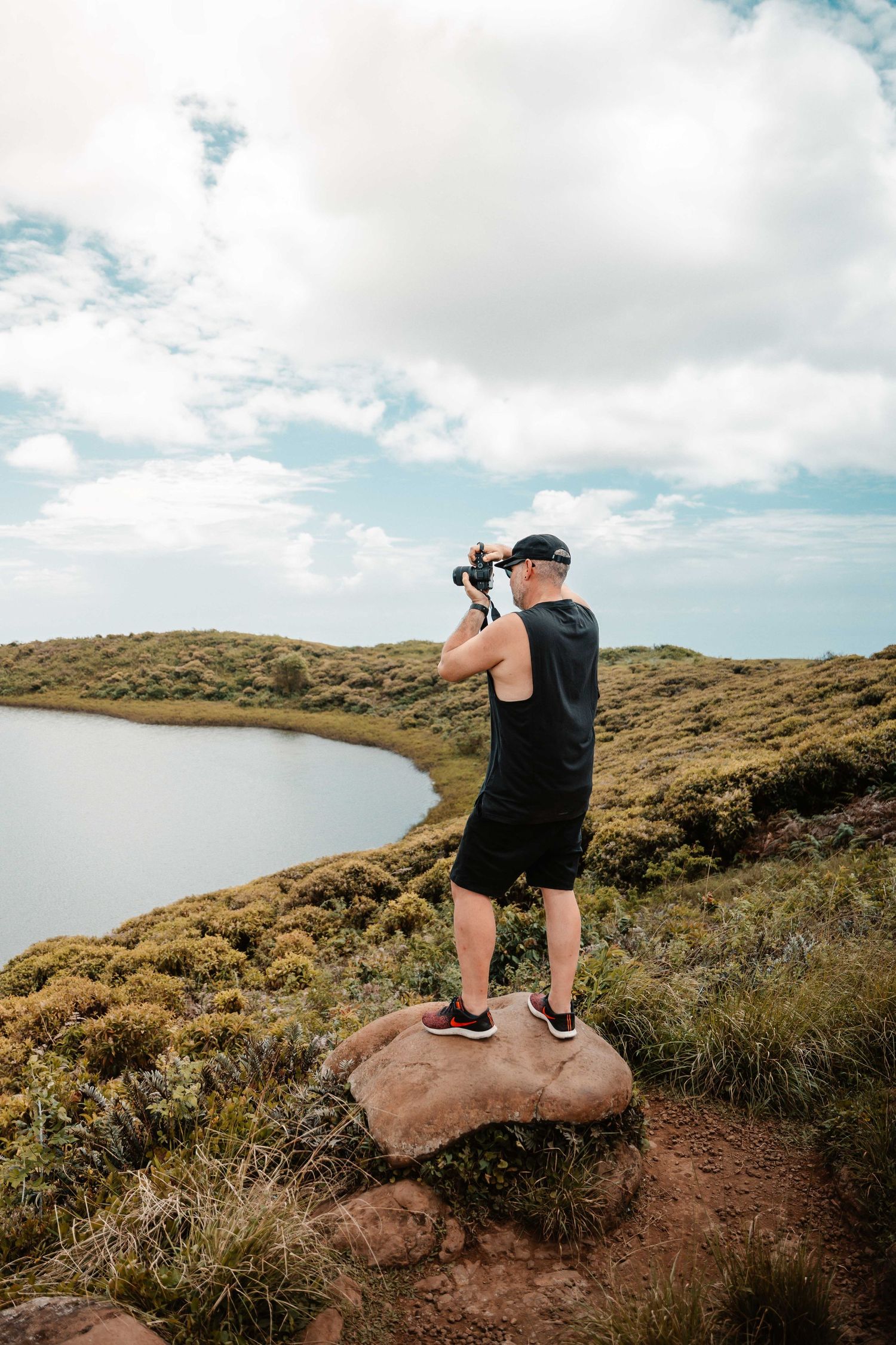 Person crouching on a rocky outcrop overlooking coastal landscape with cloudy sky.