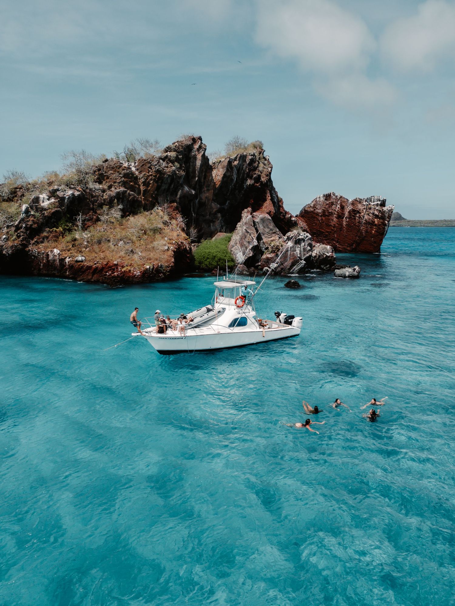 A white boat anchored near rocky outcrops in crystal clear turquoise waters with swimmers nearby.