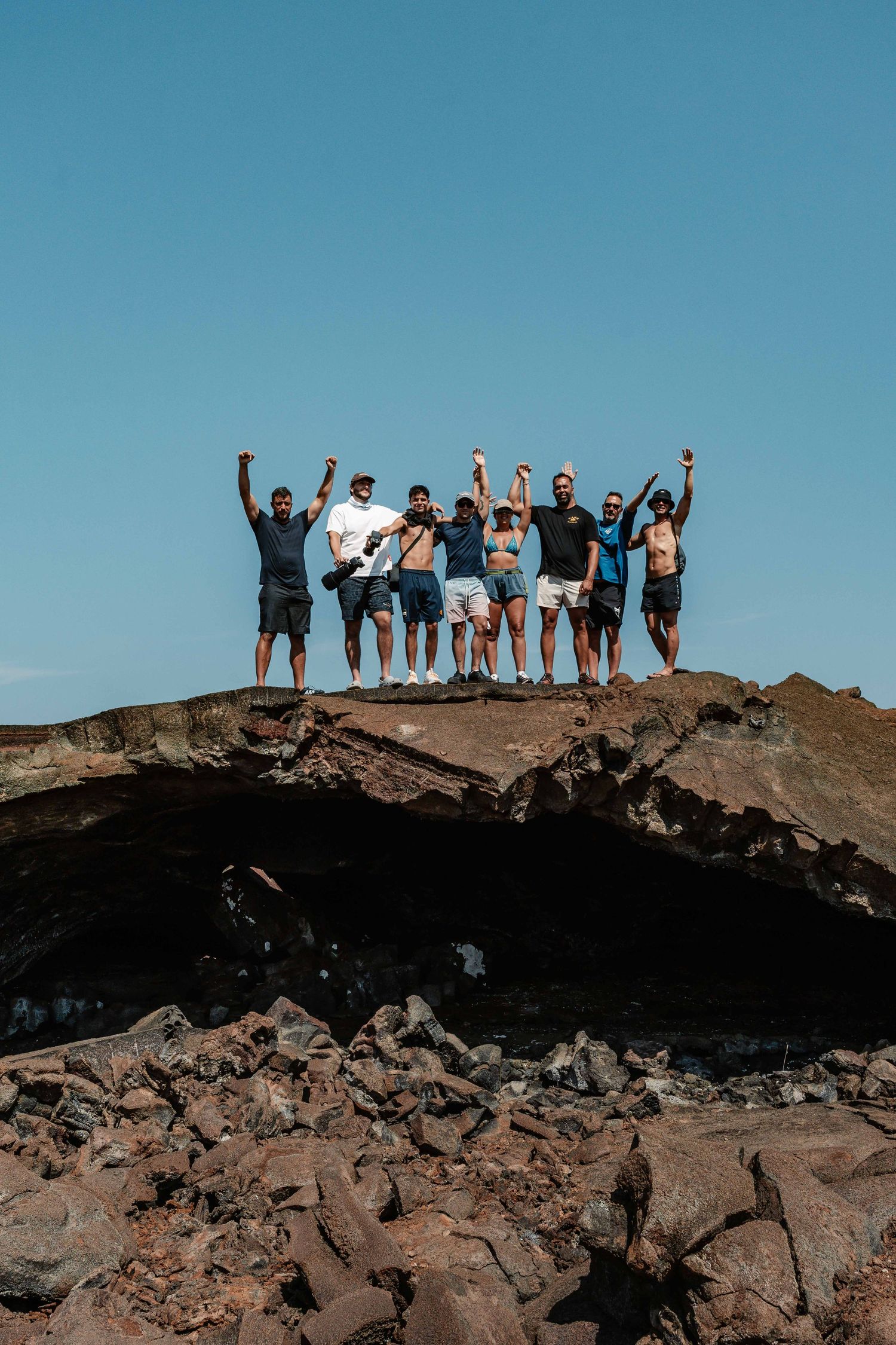 Group of people celebrating with raised arms on rocky mountain summit against blue sky.
