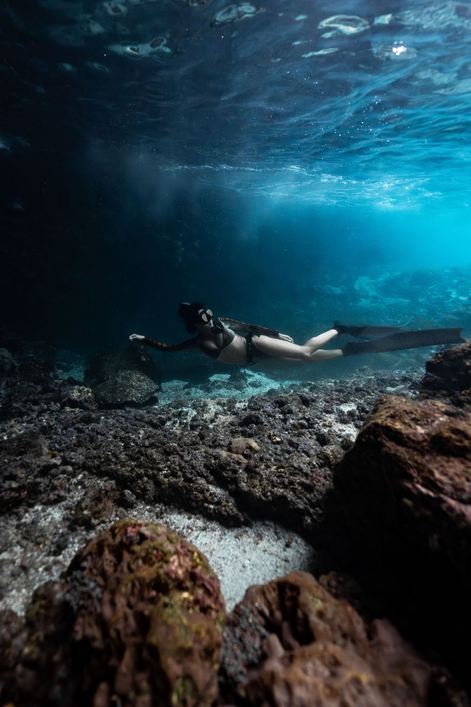 A freediver glides gracefully near the rocky ocean floor in crystal-clear blue water, illuminated by light filtering from above.