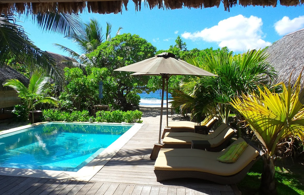A serene infinity pool with lounge chairs overlooks a tropical lagoon surrounded by lush palm trees and clear blue skies.