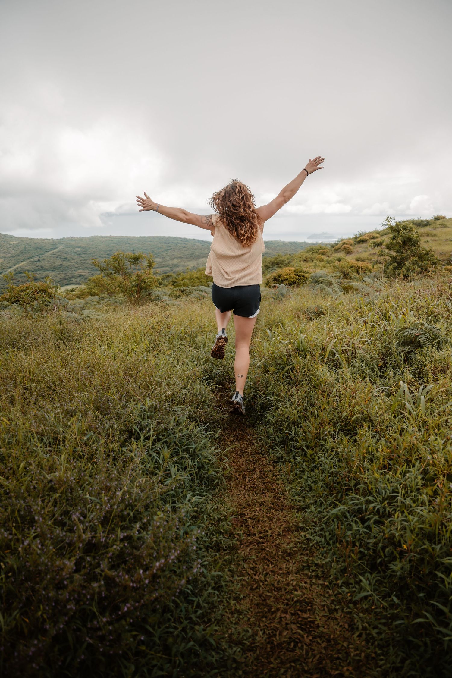 Woman with arms outstretched running on a nature trail through green fields with misty mountains in the background.