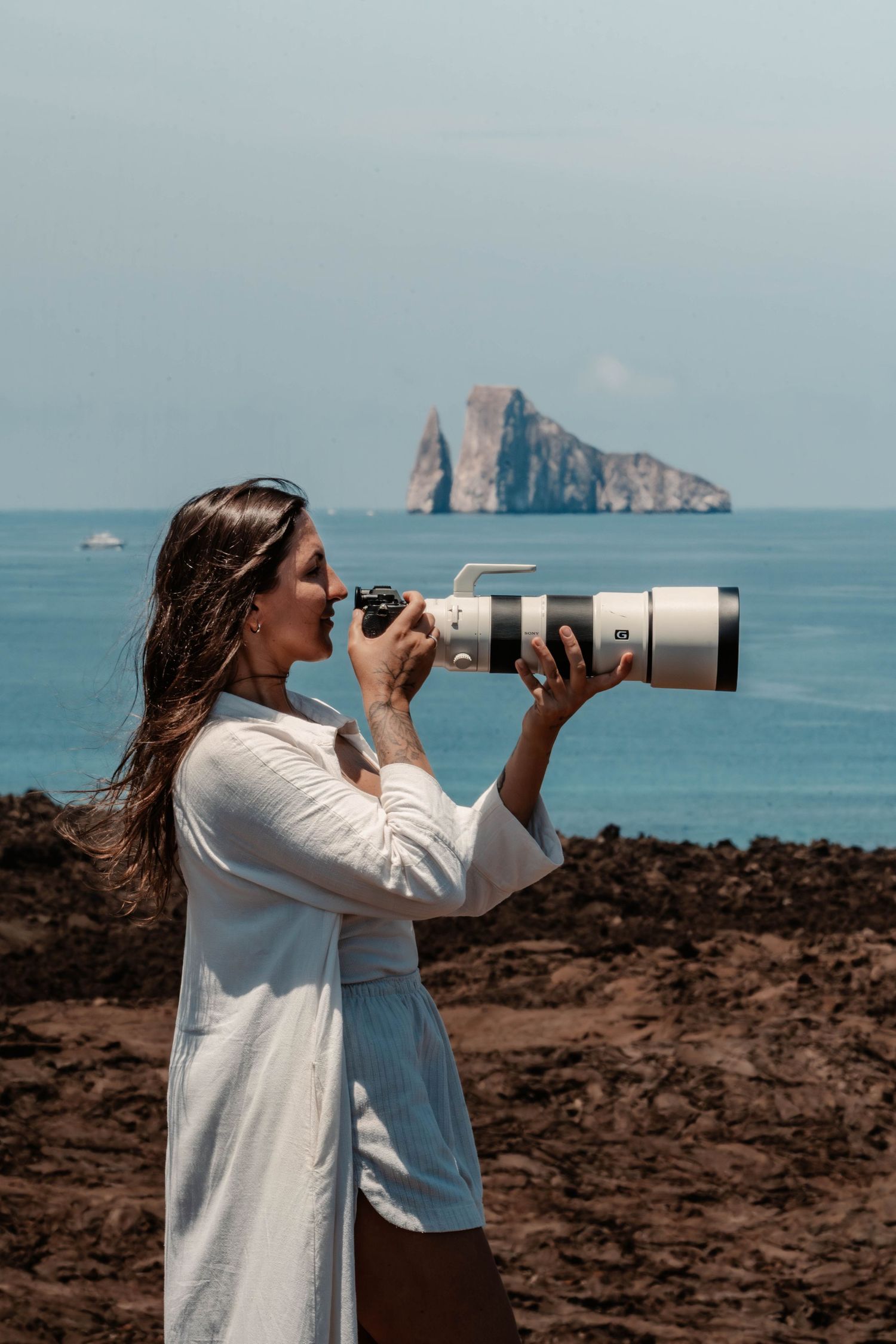 Photographer with telephoto lens capturing coastal scenery from rocky shoreline with ocean and islands in background.