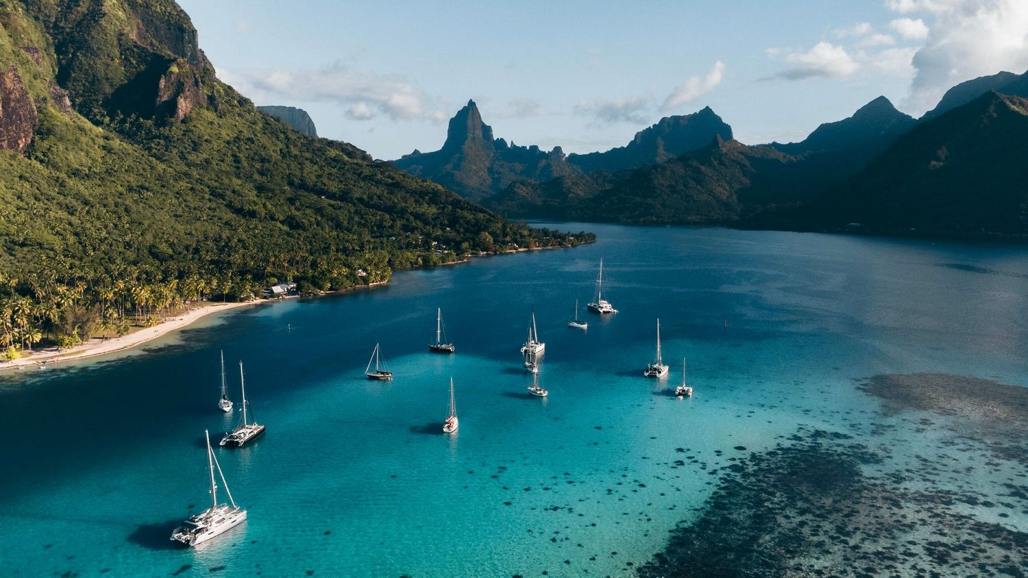 Panoramic view of tropical bay with sailboats anchored near mountainous coastline.