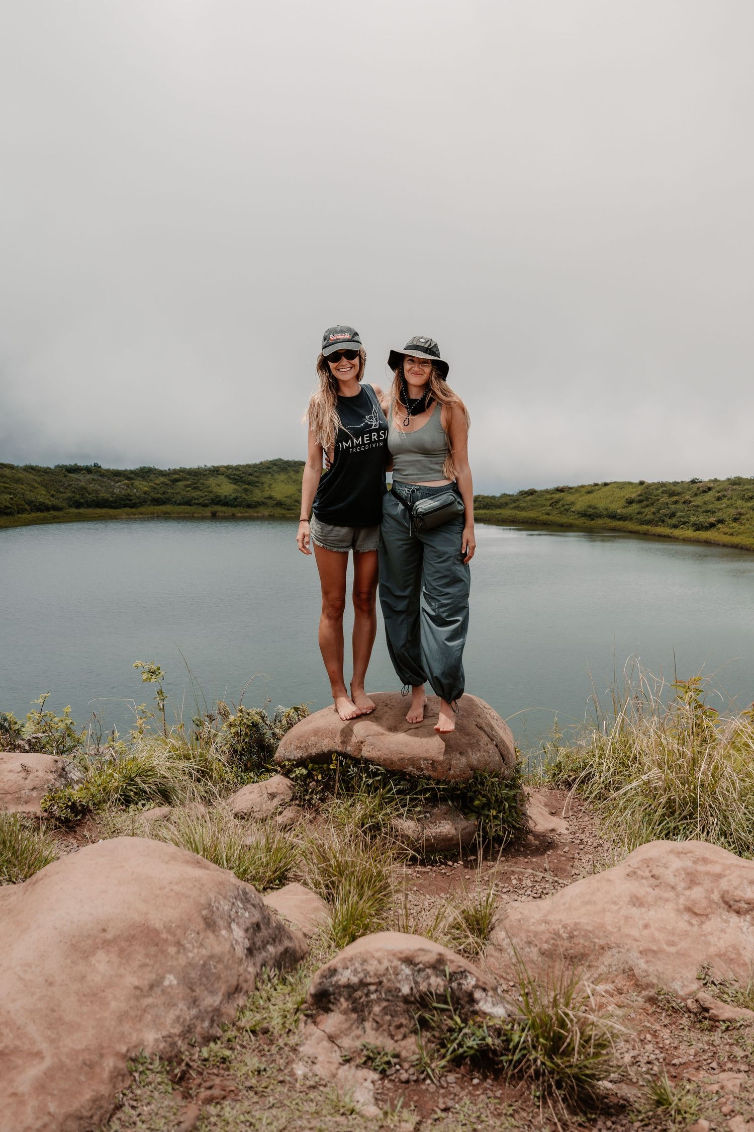 Two women standing on a rock near a scenic lake surrounded by green hills under an overcast sky.