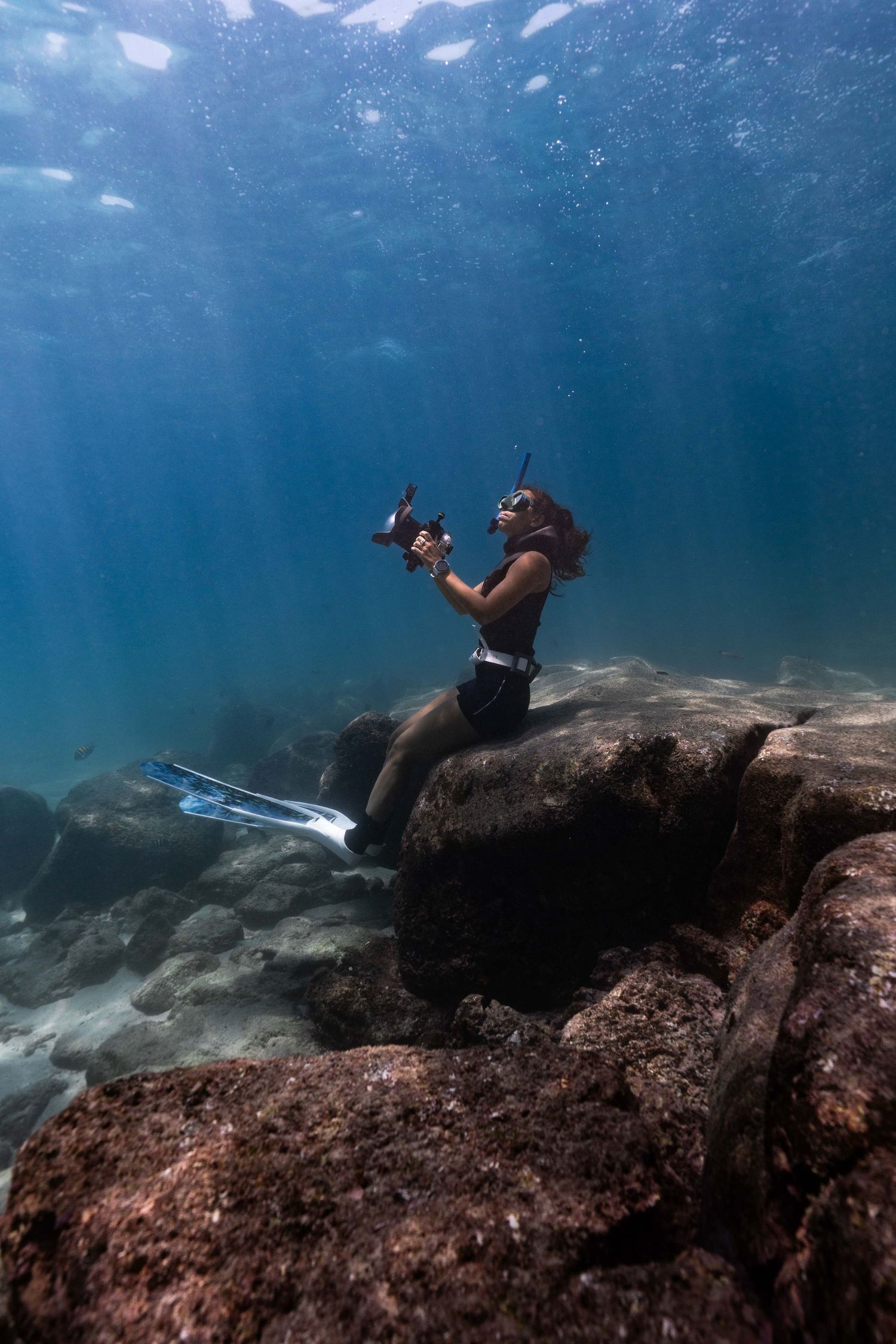 Scuba diver in wetsuit sitting on underwater rocks against deep blue ocean background with snorkel gear.