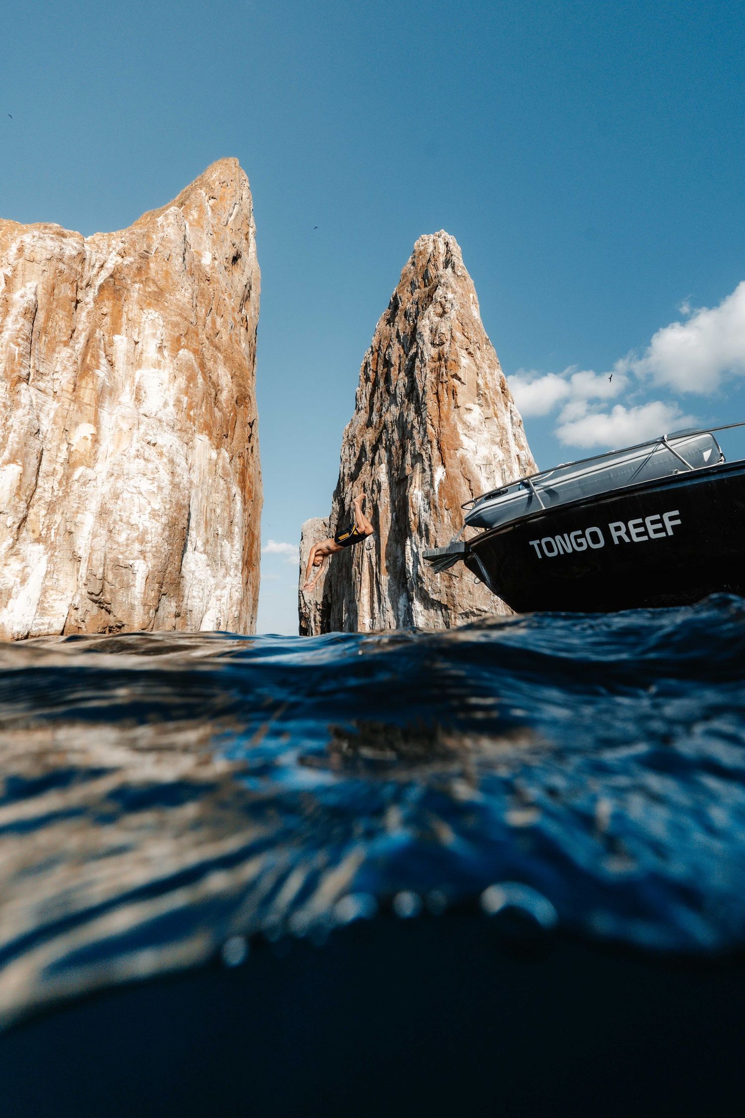 Split-view underwater photo of a rocky cliff formation and boat called Tongo Reef against blue sky.