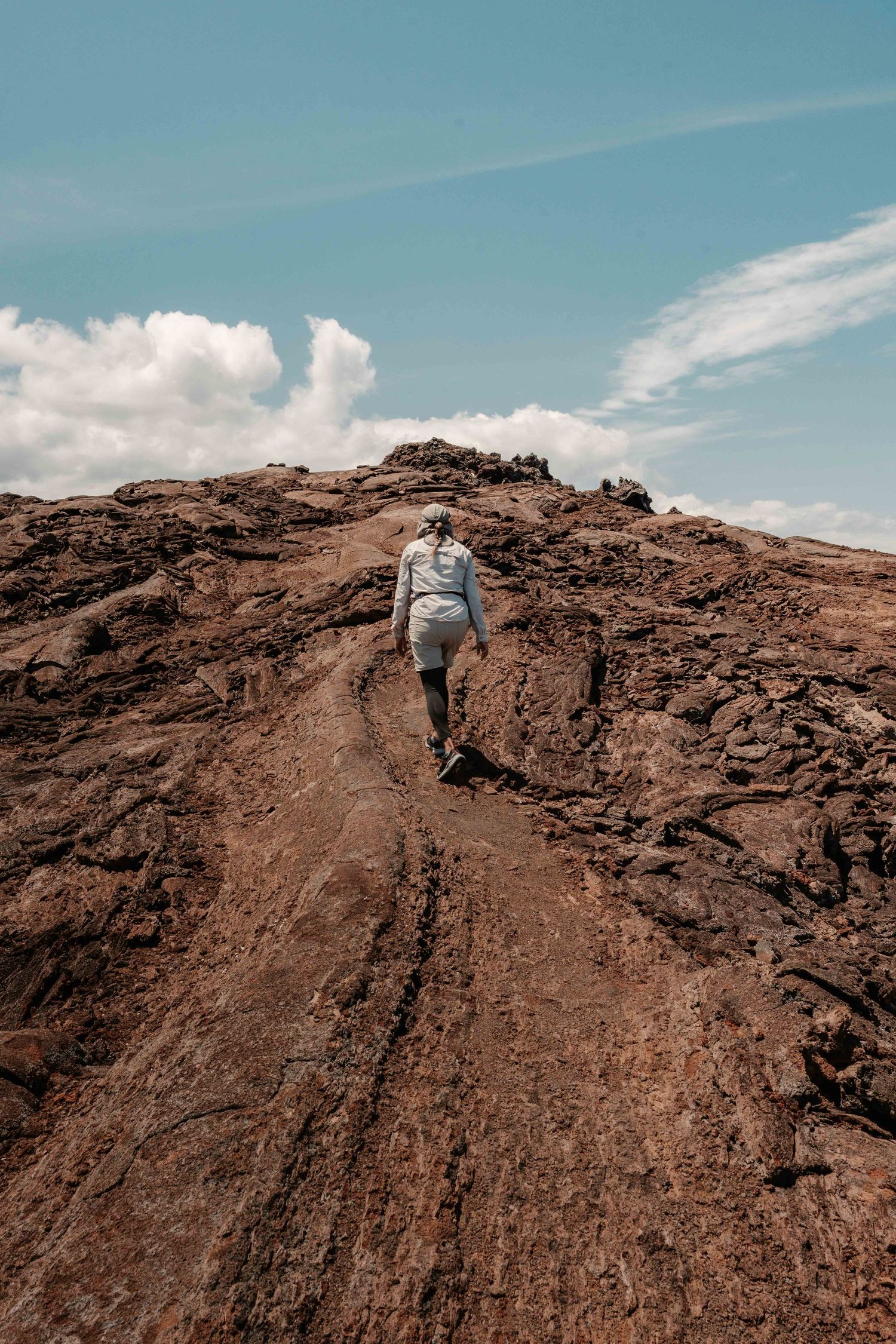 Hikers ascending a steep rocky volcanic terrain under bright blue skies with wispy clouds.