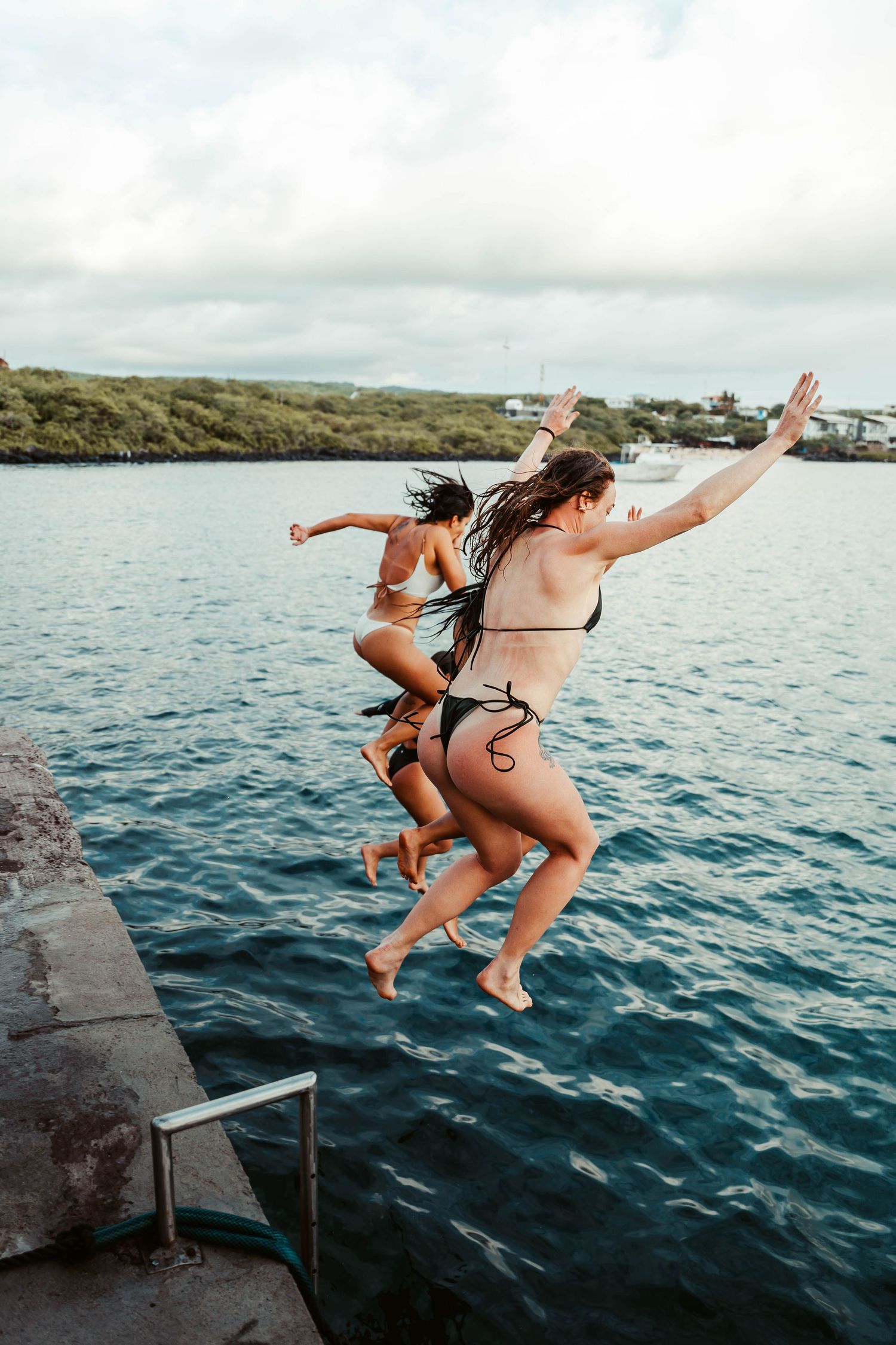 Two friends in swimsuits leap joyfully from a rocky ledge into blue ocean waters on a sunny day.