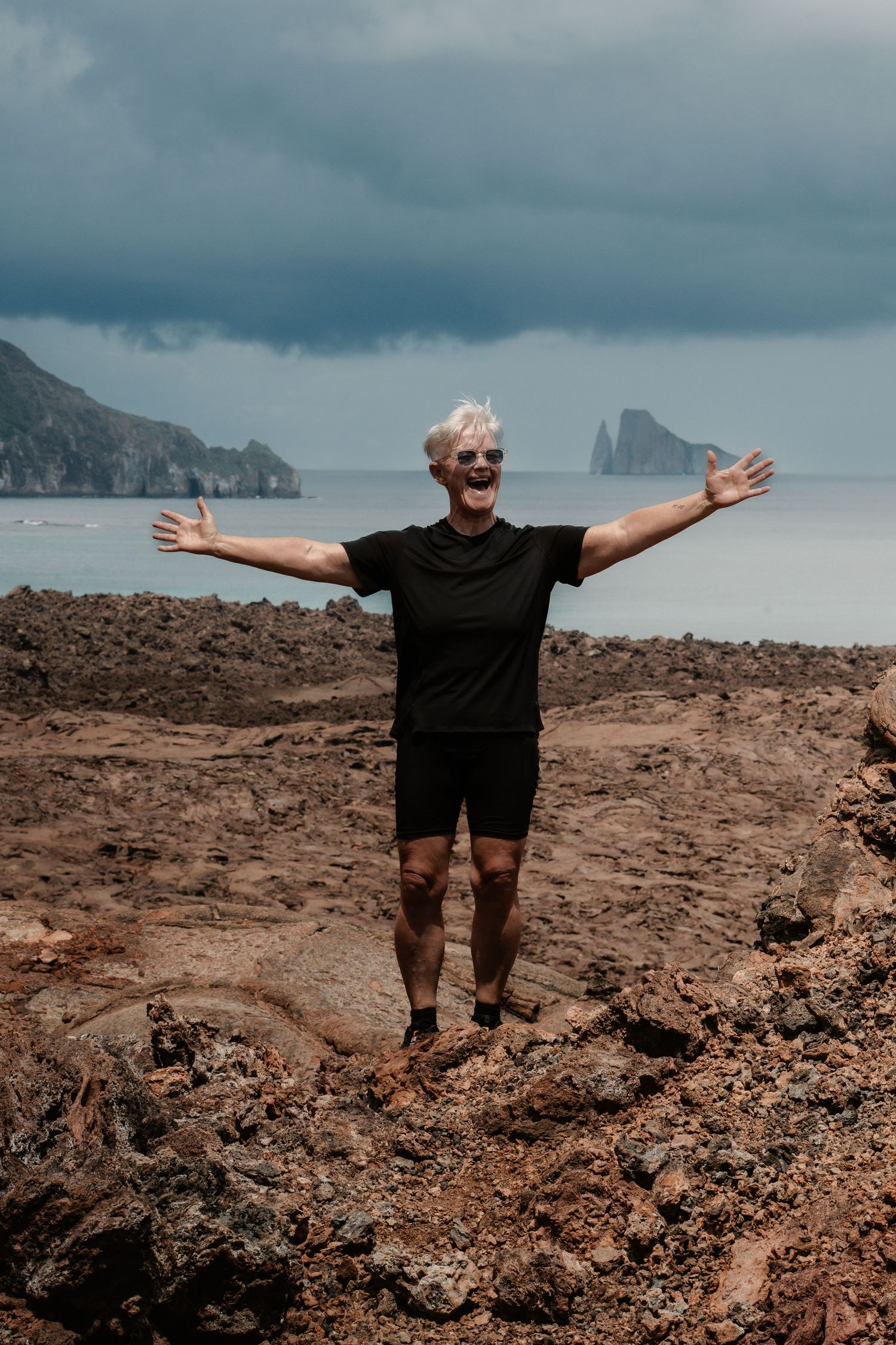 A joyful man with white hair spreads his arms wide on rocky volcanic terrain with stormy sea and cliffs behind him.