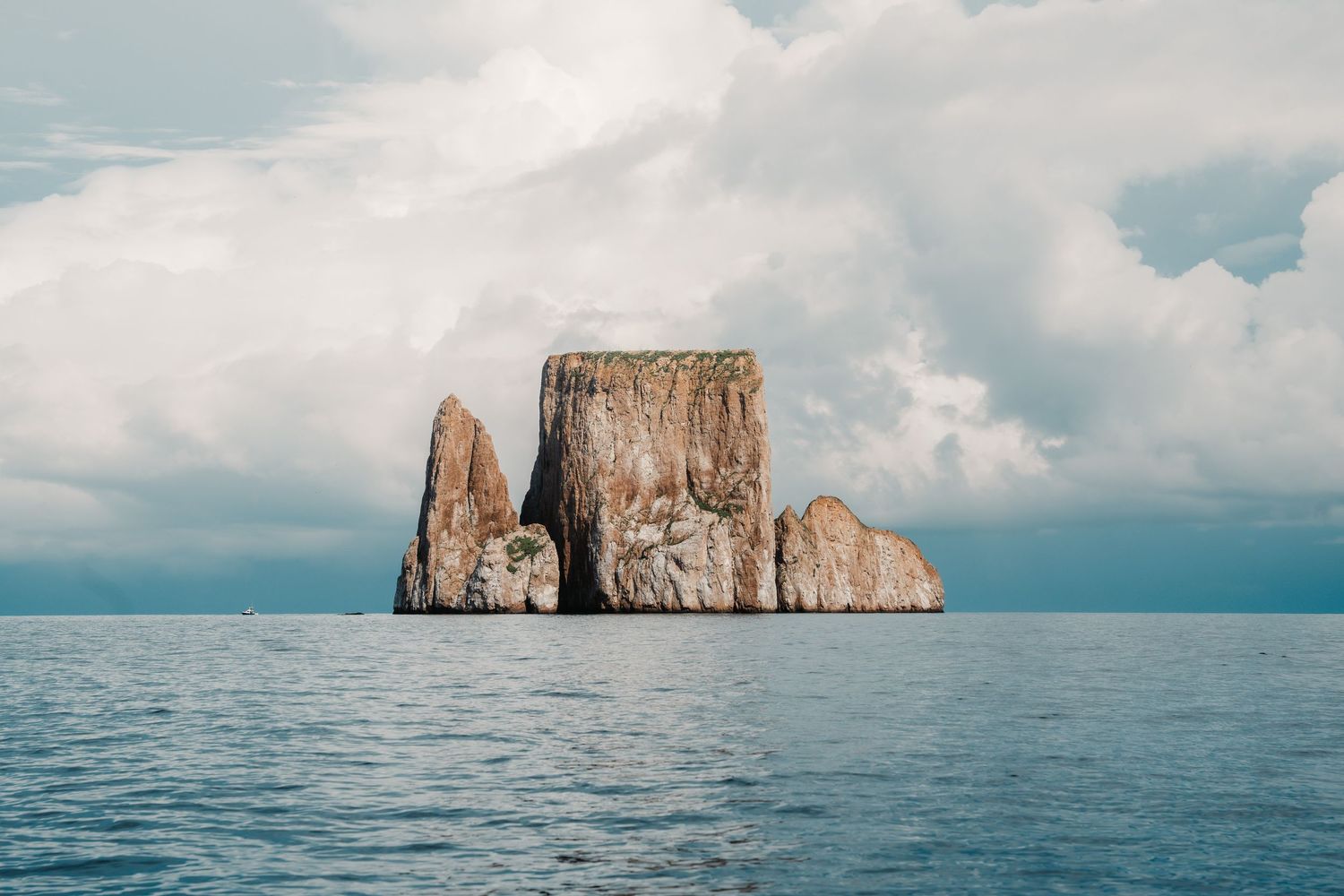 A dramatic rocky sea stack rises from calm blue ocean waters beneath a cloudy sky in the Galapagos Islands.