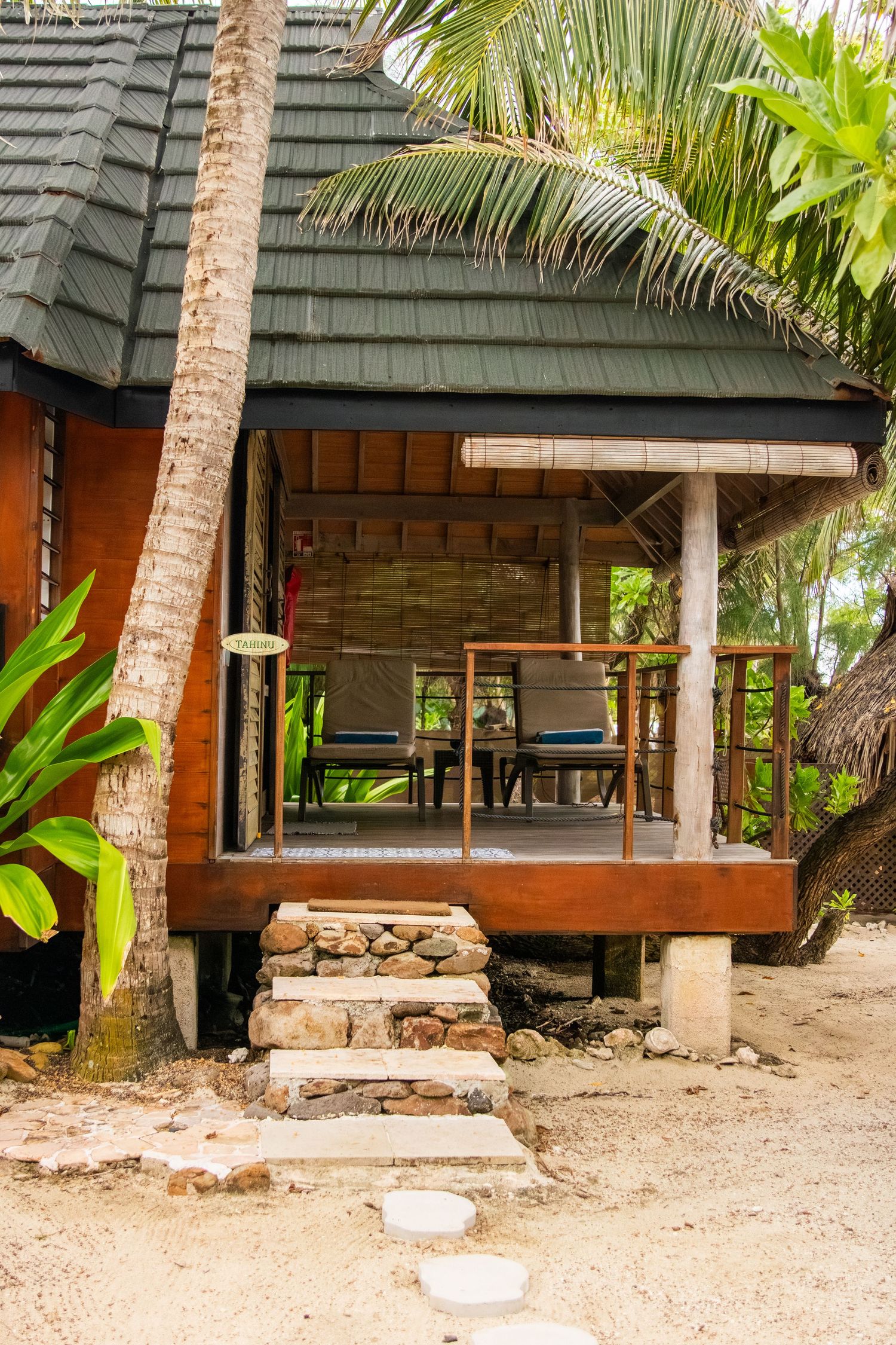 Rustic beach hut with thatched roof and wooden deck surrounded by tropical palm trees and sandy ground.