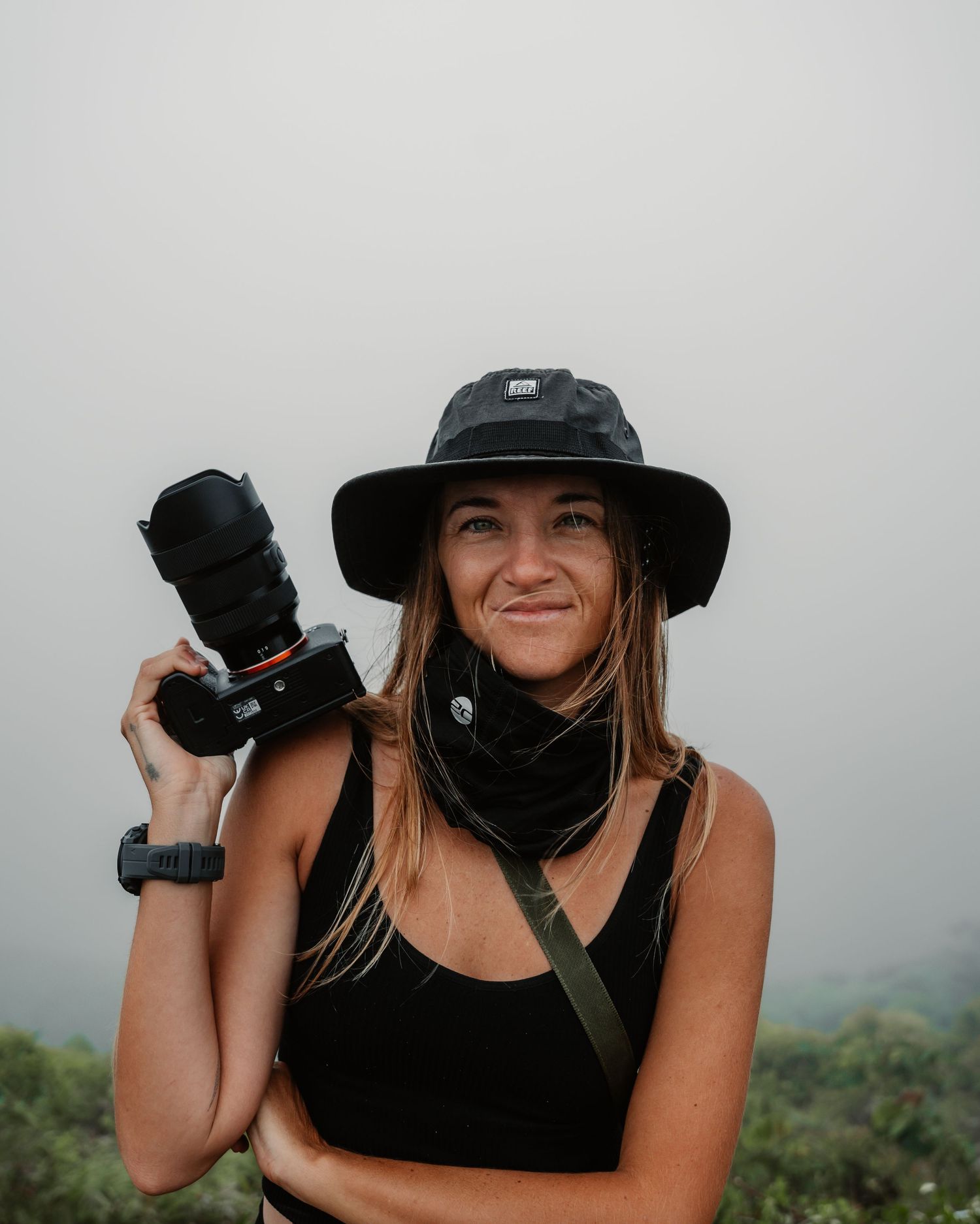 A photographer in a black tank top and hat takes photos in a misty mountain landscape.