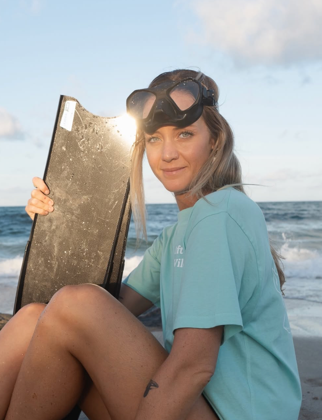 Someone in a turquoise shirt sits on a beach reading a book as waves crash in the background during sunset.