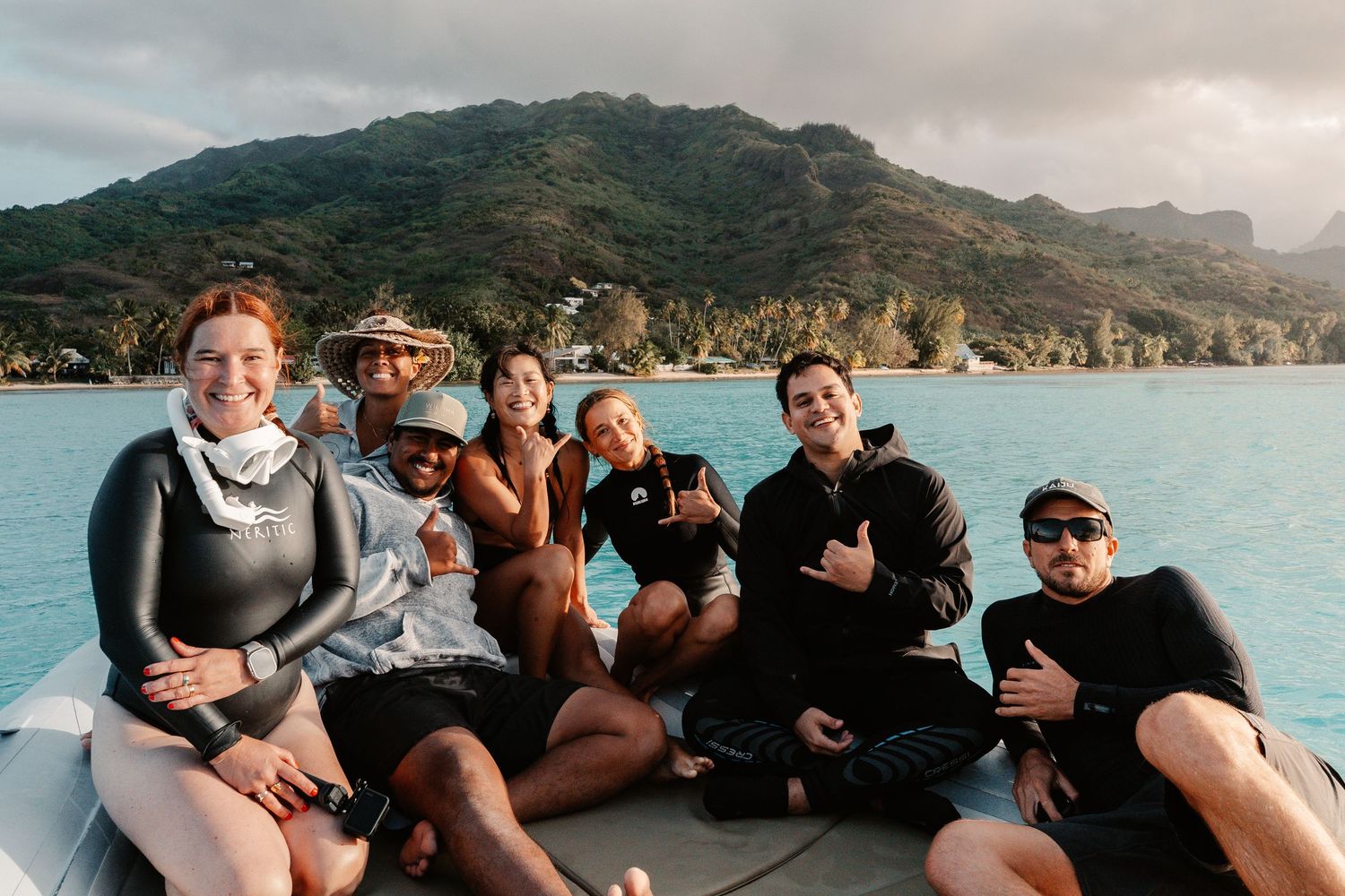 Group of seven divers smiling on a boat with tropical mountains and turquoise waters in the background.