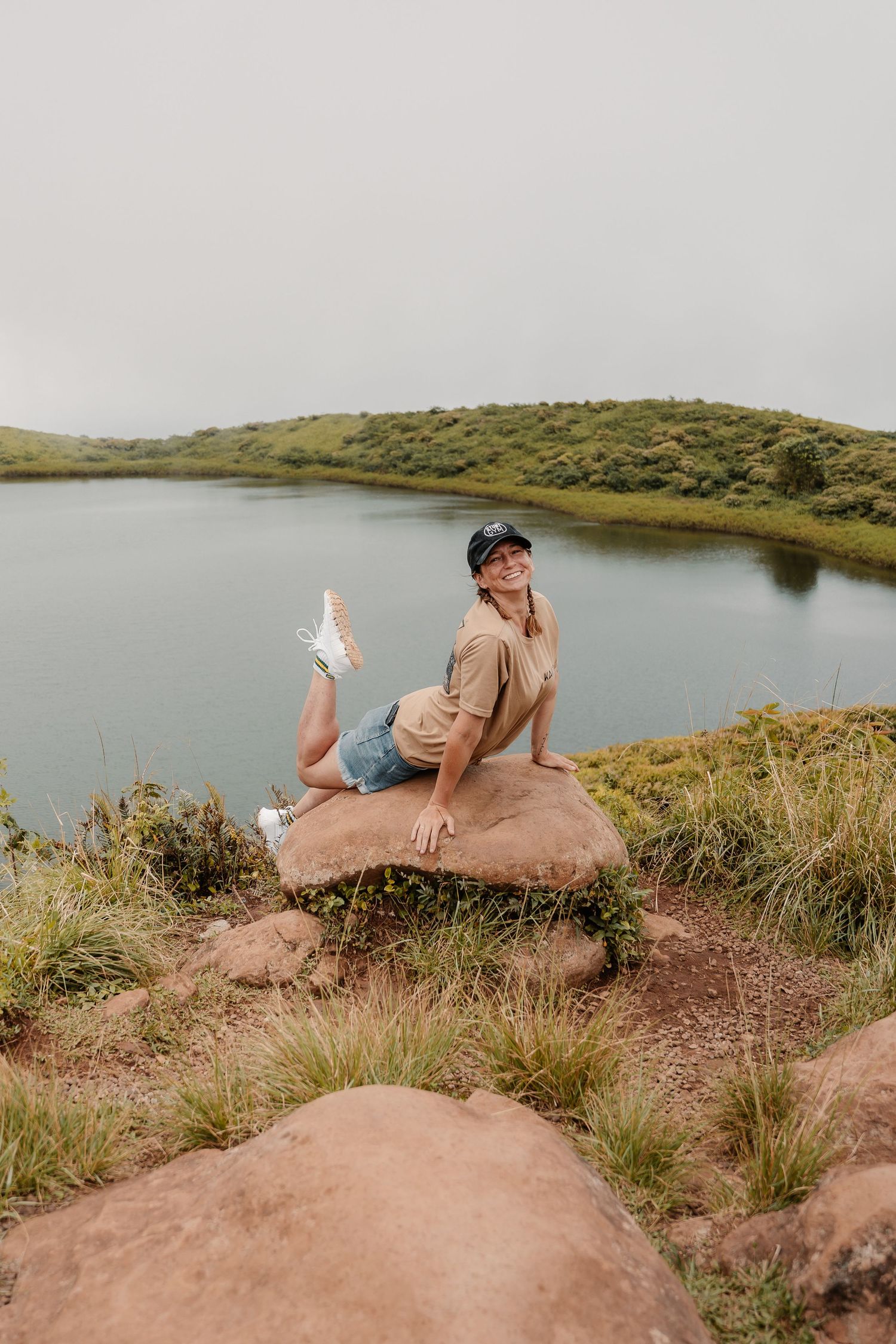 Woman posing on a rock by a scenic lake with lush green hills under an overcast sky.