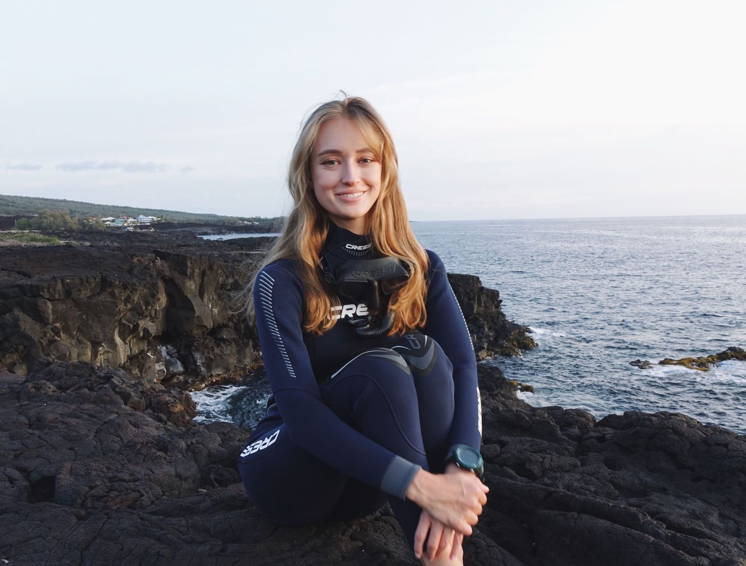 A person in a blue wetsuit sits on dark volcanic rocks along a scenic ocean coastline.