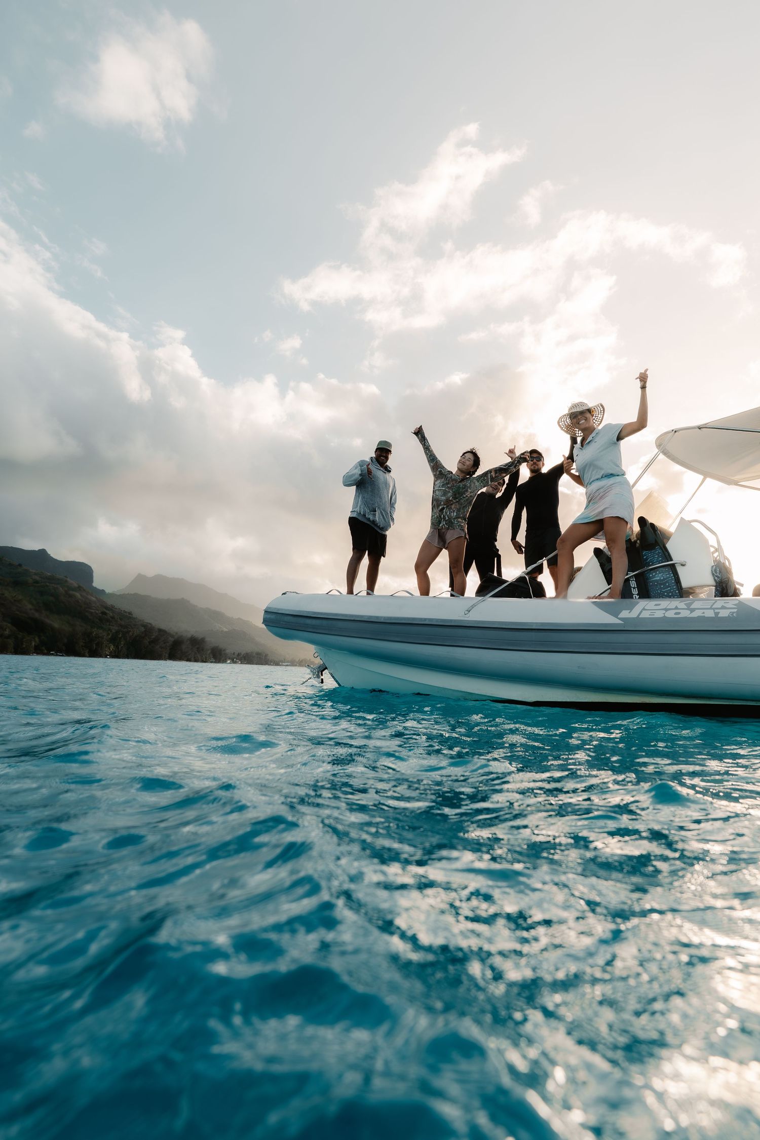Friends celebrating on a white inflatable boat in turquoise blue ocean waters with mountains in the background.