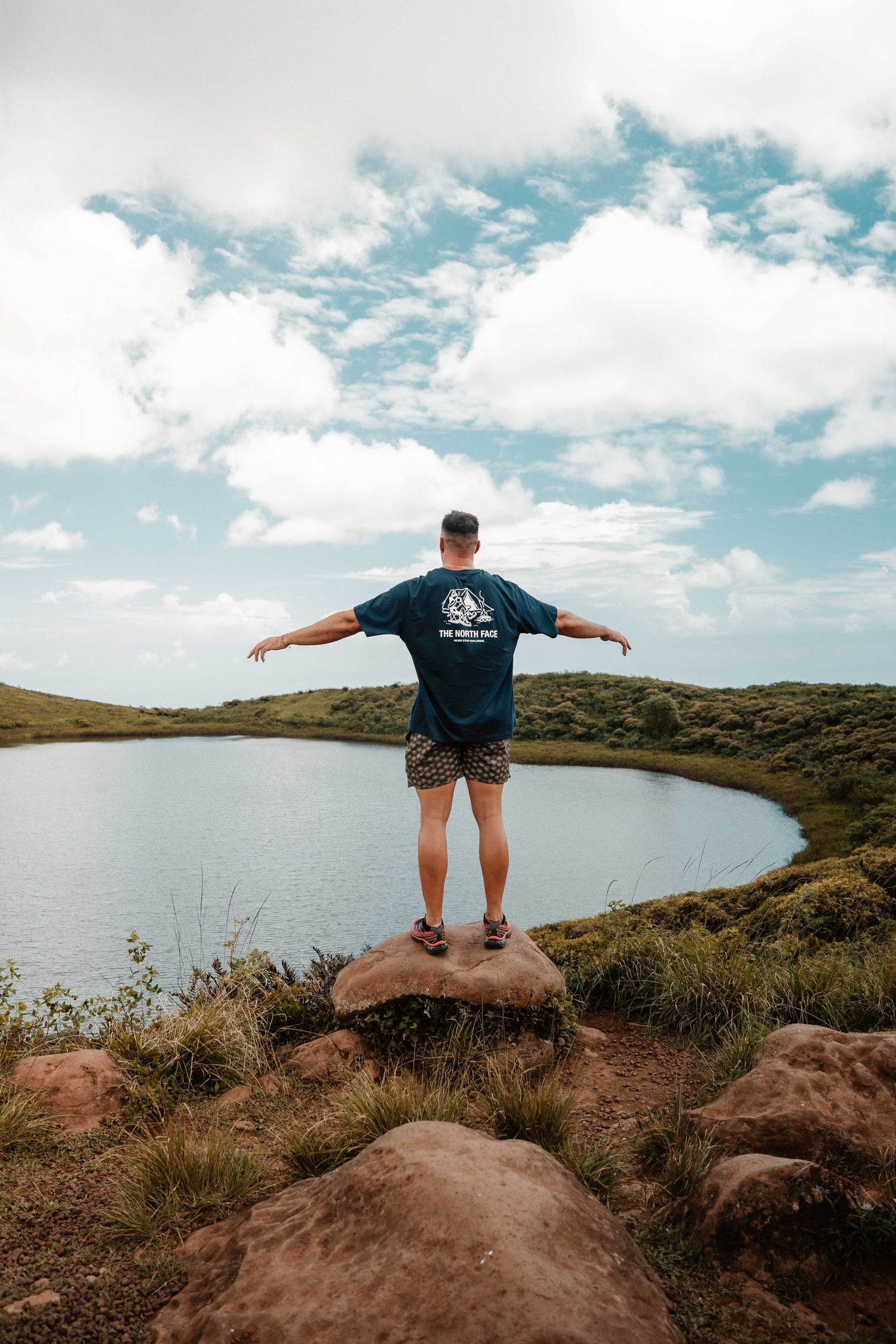 Person stands with arms outstretched on rocky overlook above a curved lake under blue sky with white clouds.