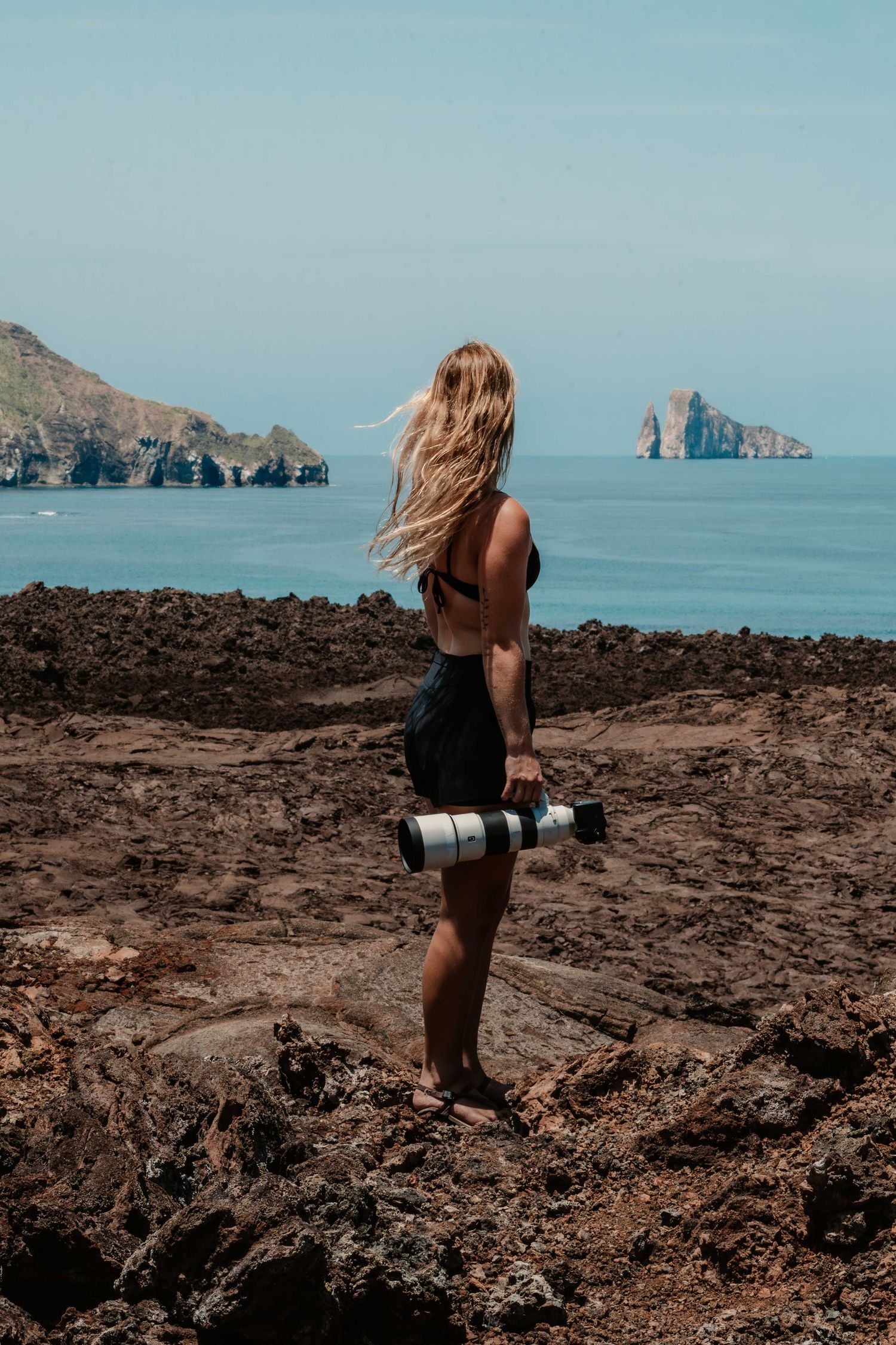 Photographer with long hair stands on rocky cliff overlooking blue ocean and dramatic rock formations in distance.