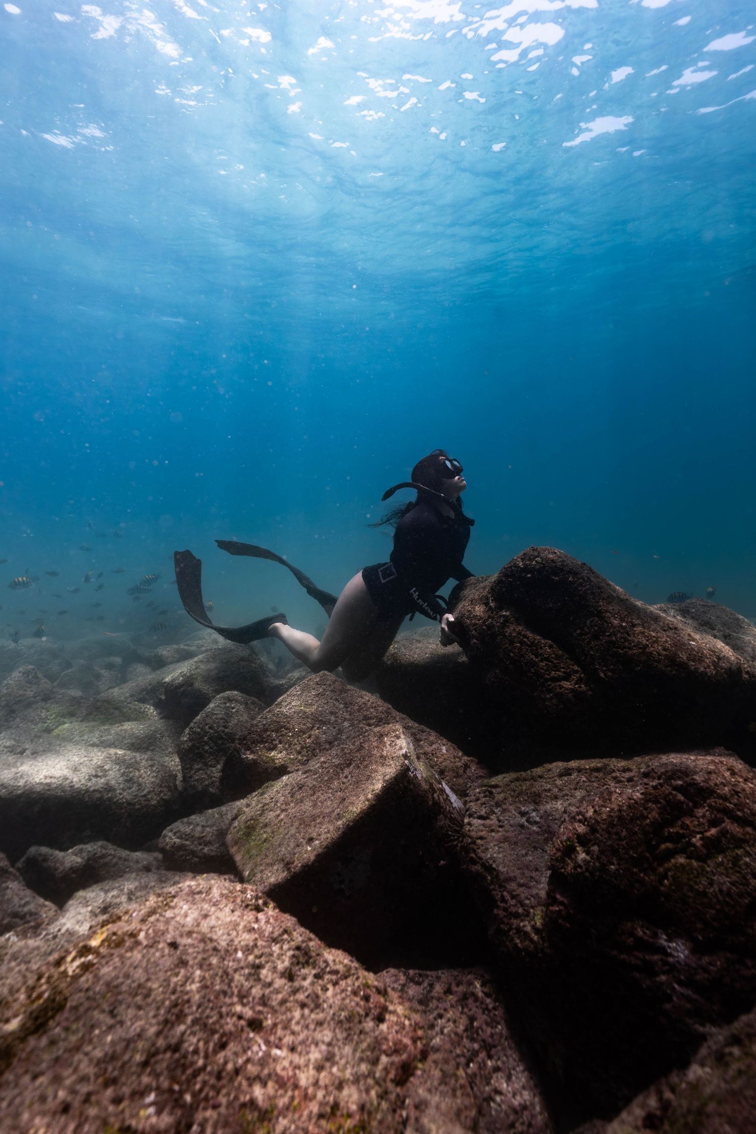 Scuba divers exploring underwater rock formations in crystal clear blue waters with sunlight filtering from above.