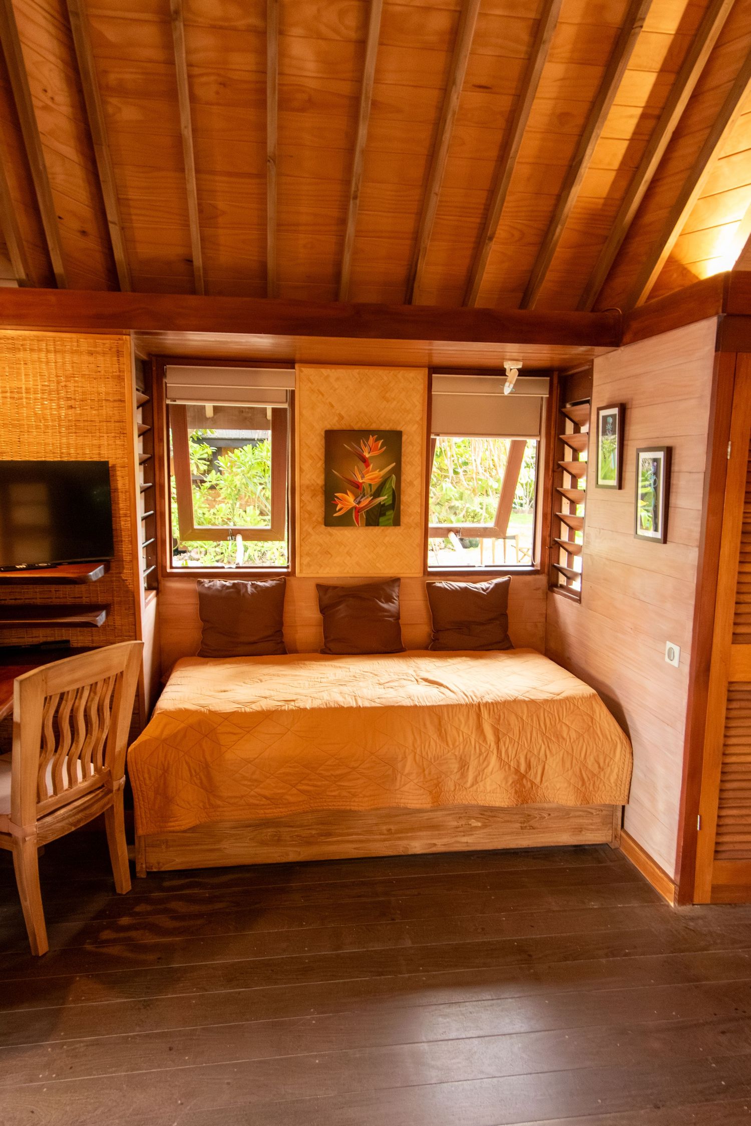 Cozy wooden bungalow bedroom with warm tones, a double bed, tropical artwork, shuttered windows, and a flat-screen TV.