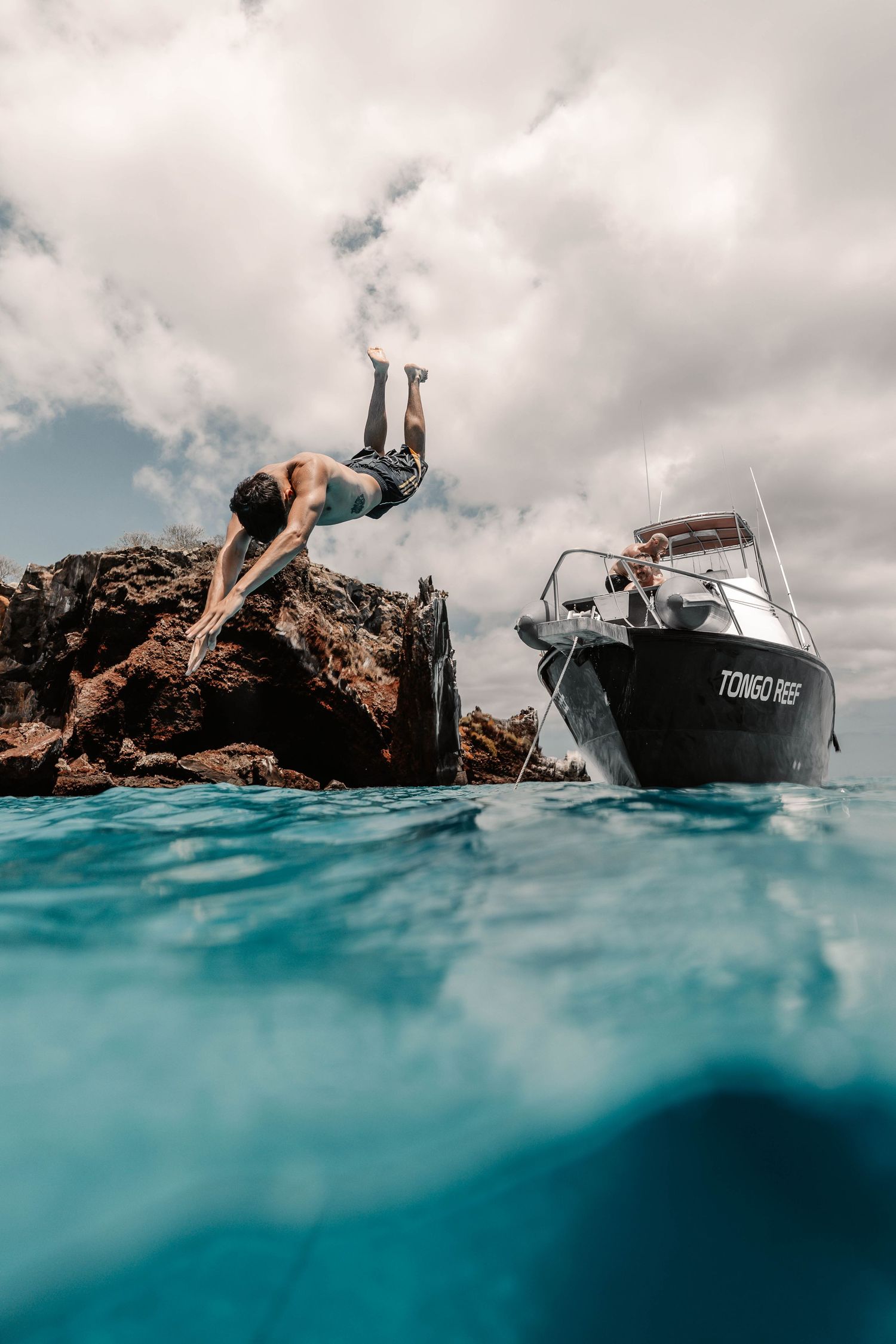 Someone diving off rocky cliffs into turquoise waters near a boat on a cloudy day.