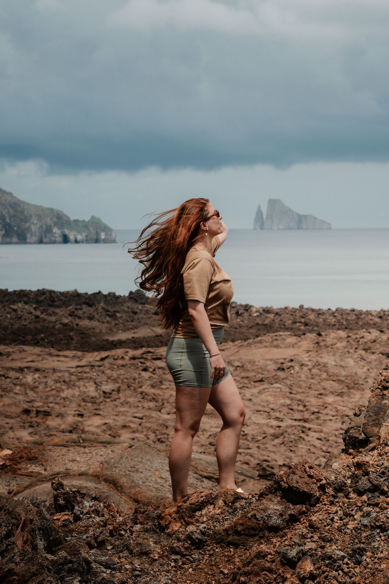 Red-haired woman in khaki outfit poses on rocky coastal landscape with dramatic sea stacks and stormy sky.