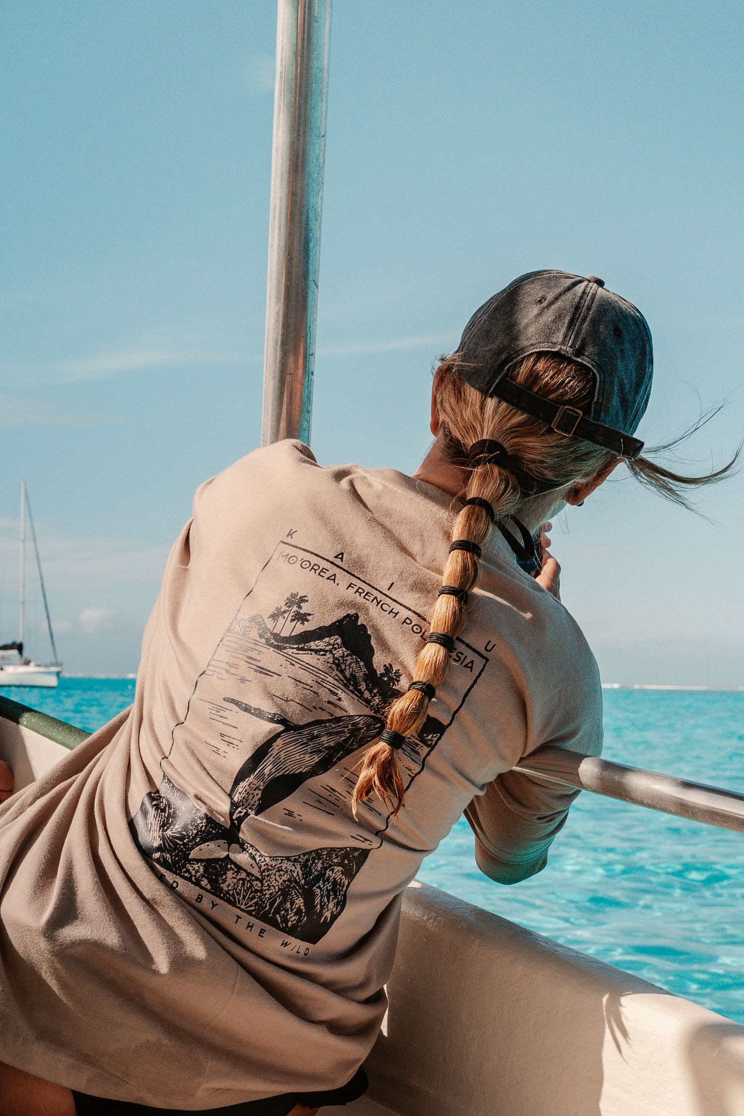 Person with braided hair in cap and graphic tee sits on boat overlooking turquoise ocean under blue sky.