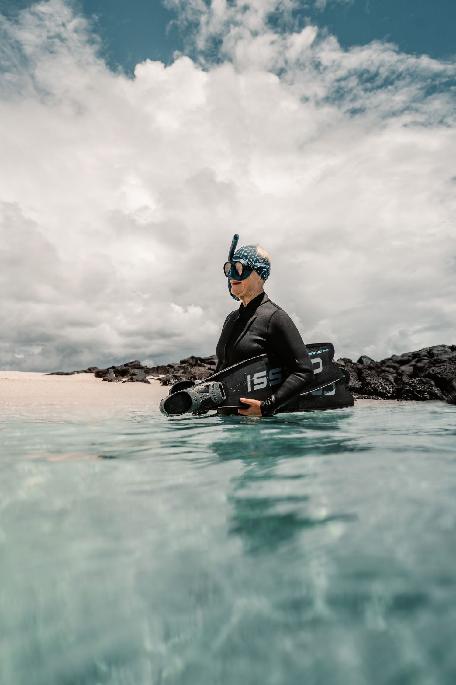 A snorkeler in a black wetsuit and mask holds fins while standing in clear shallow tropical waters near rocky shore.