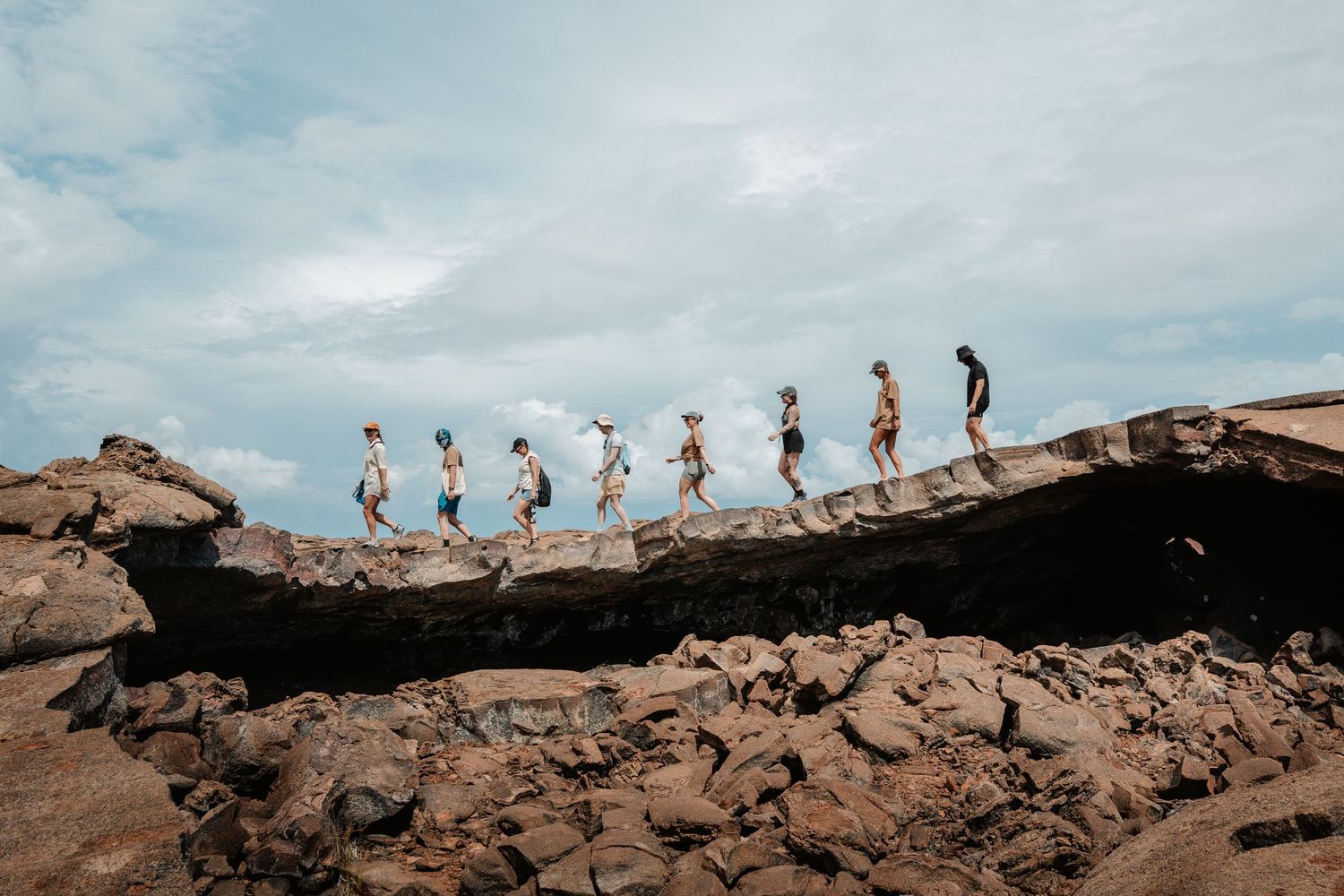A group of people walking single file across rocky coastal terrain under a cloudy sky.