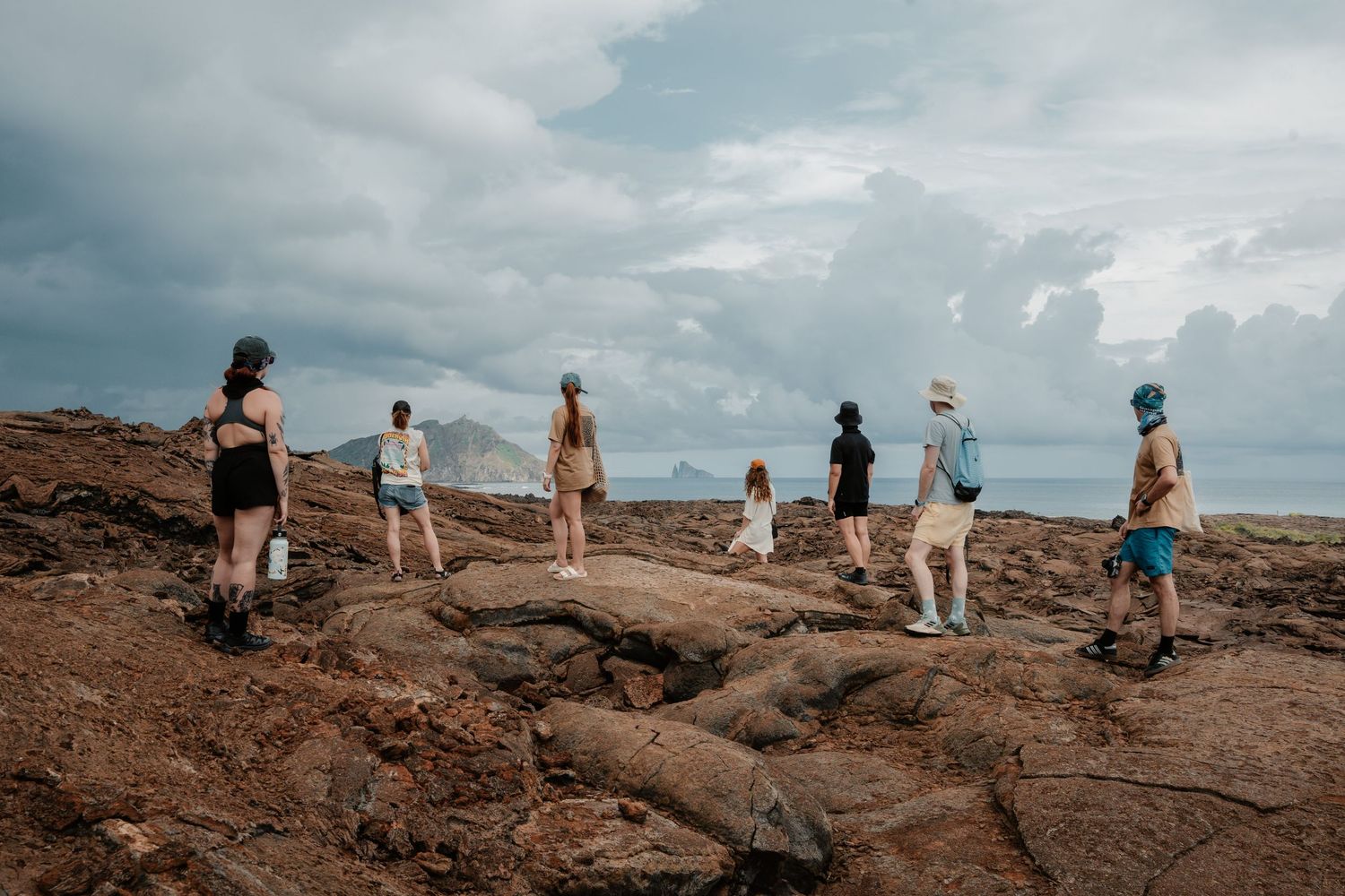 Group of hikers standing on rocky coastal terrain under dramatic cloudy skies with ocean and mountains in background.