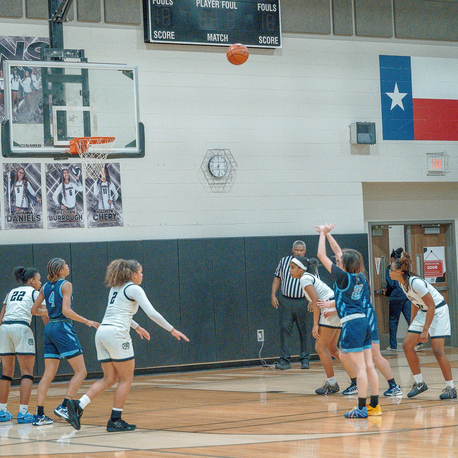 Basketball players compete in a high school gymnasium with a Texas state flag visible on the wall during a game.