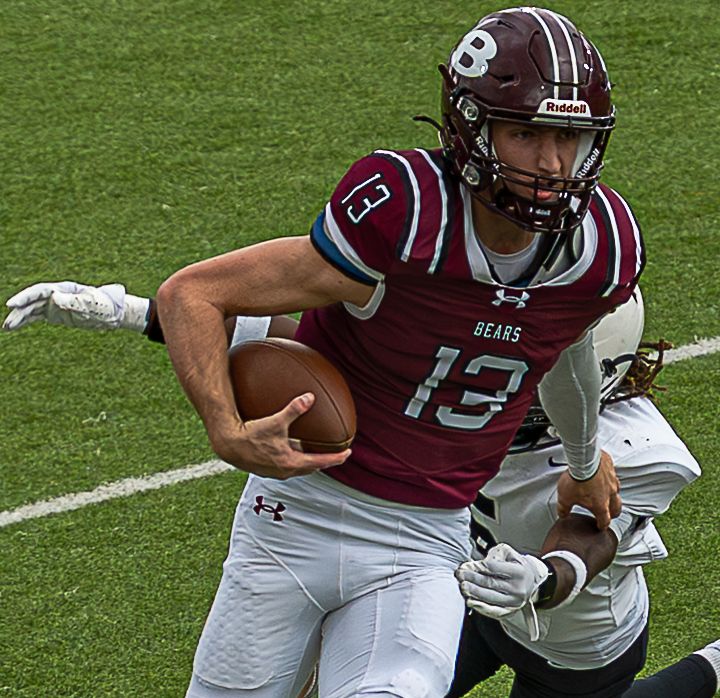 Football player in maroon and white uniform runs with the ball while being tackled during a game on grass field.