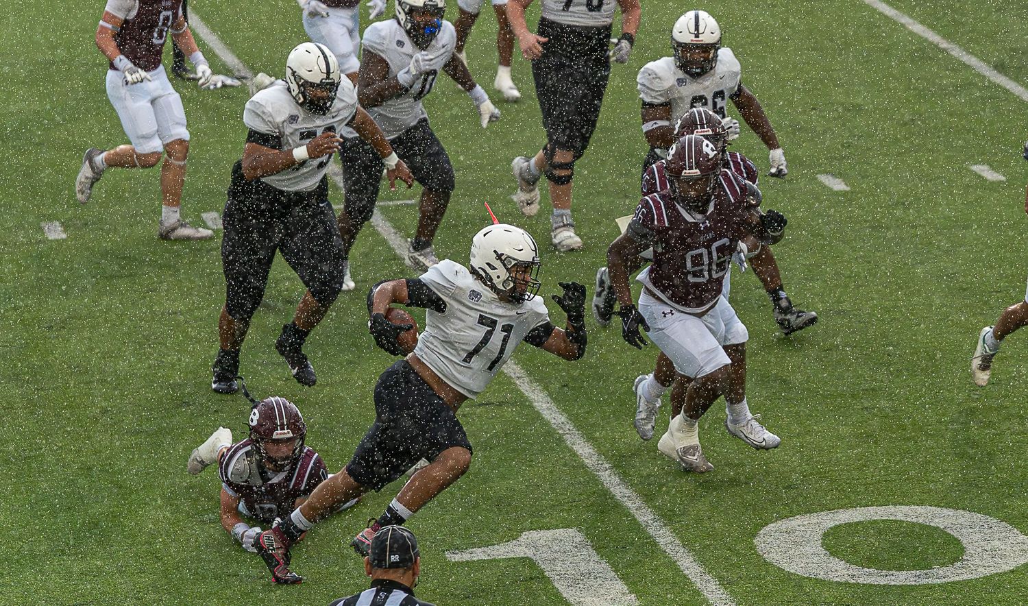 A football play in progress showing multiple players engaged in blocking and tackling on a grass field.