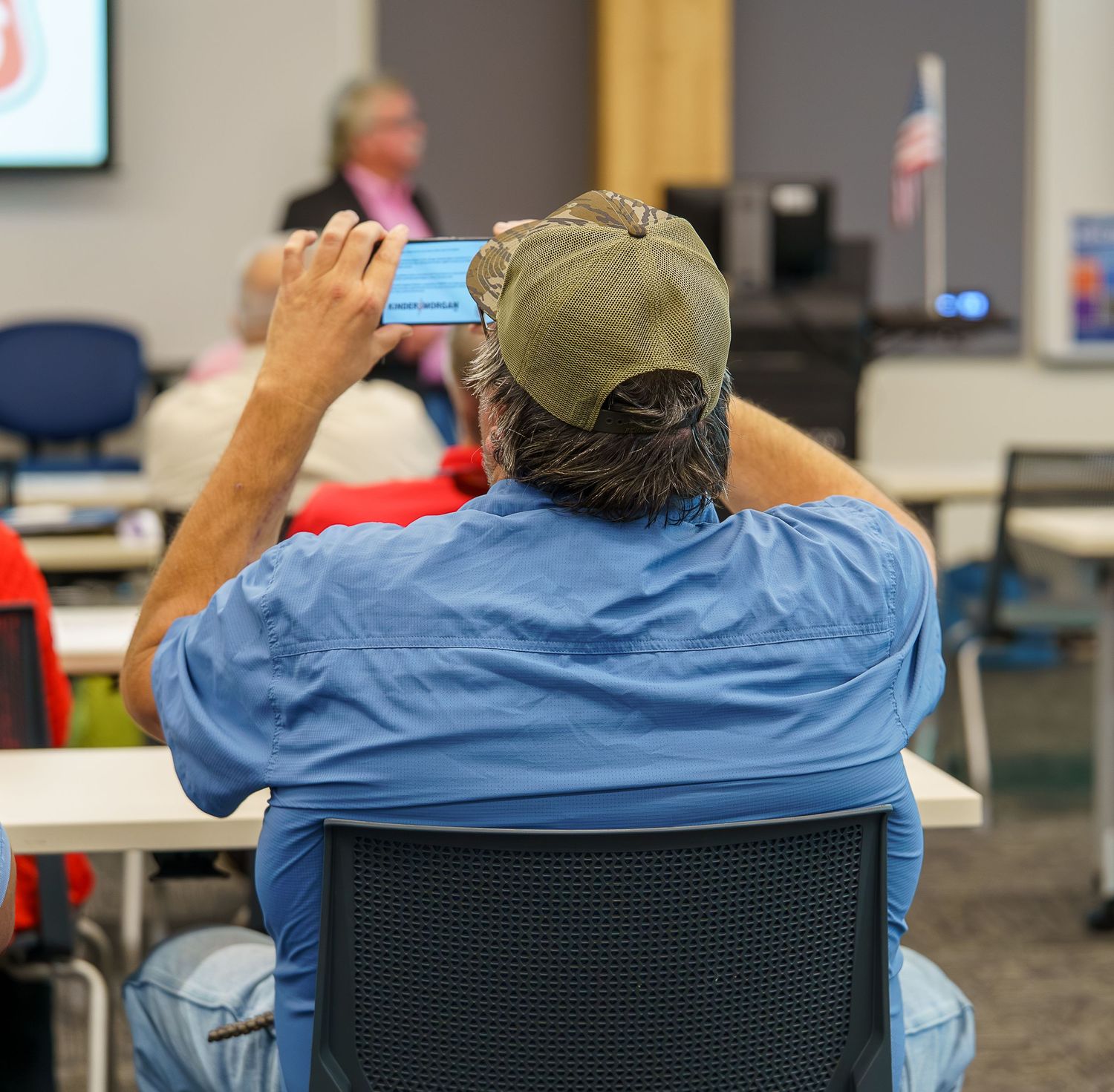 Audience member in baseball cap taking photo during presentation.