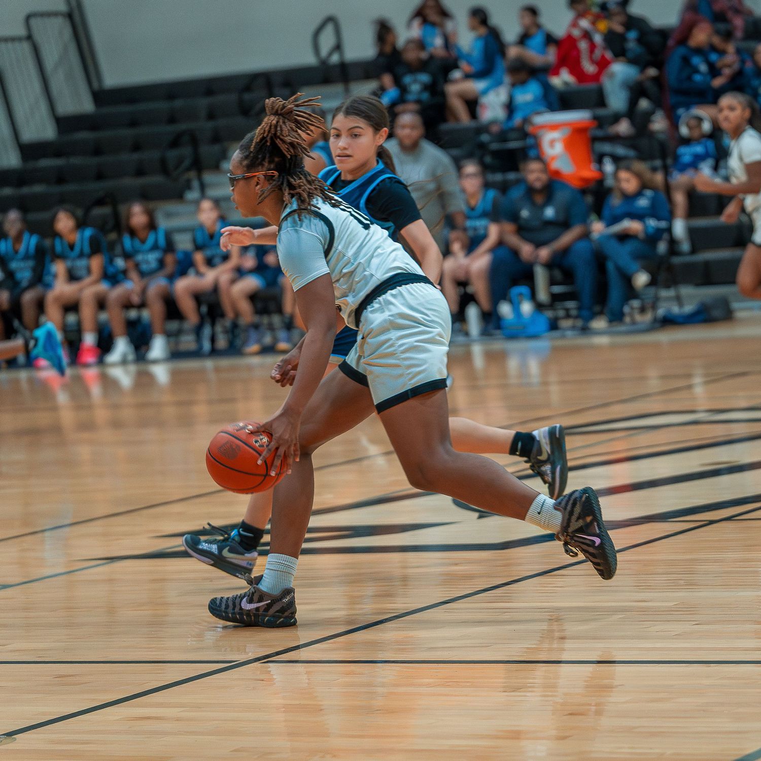Basketball player in light blue uniform dribbles down court during indoor game with spectators in background.