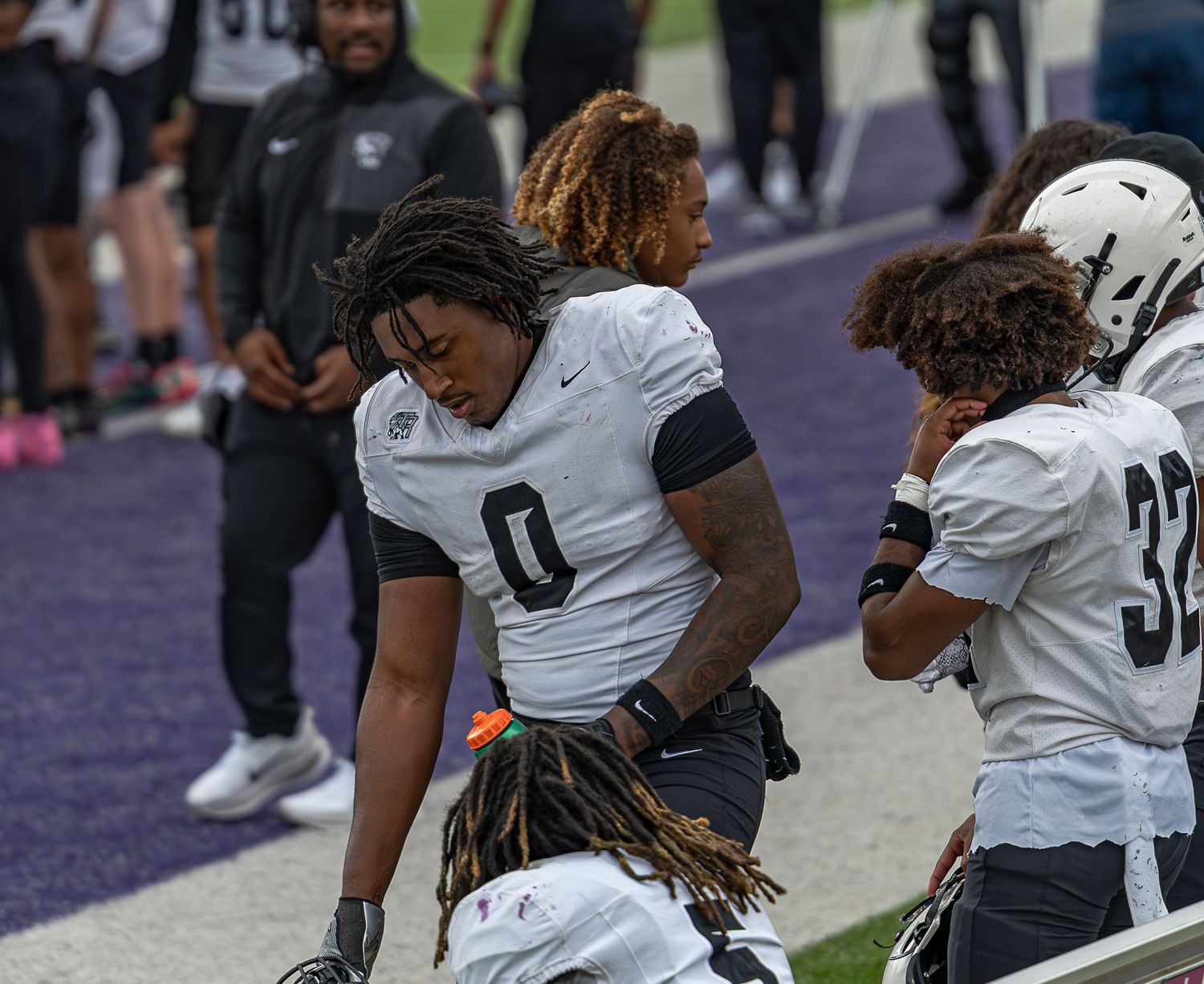 Football players in white uniforms stand on the sideline during a game.