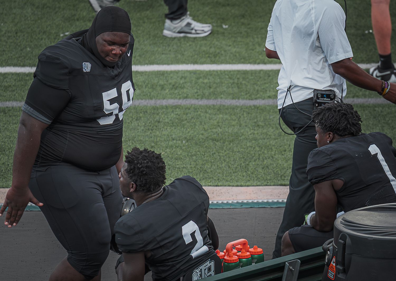 Football players in dark uniforms rest on sideline benches during a game or practice session.