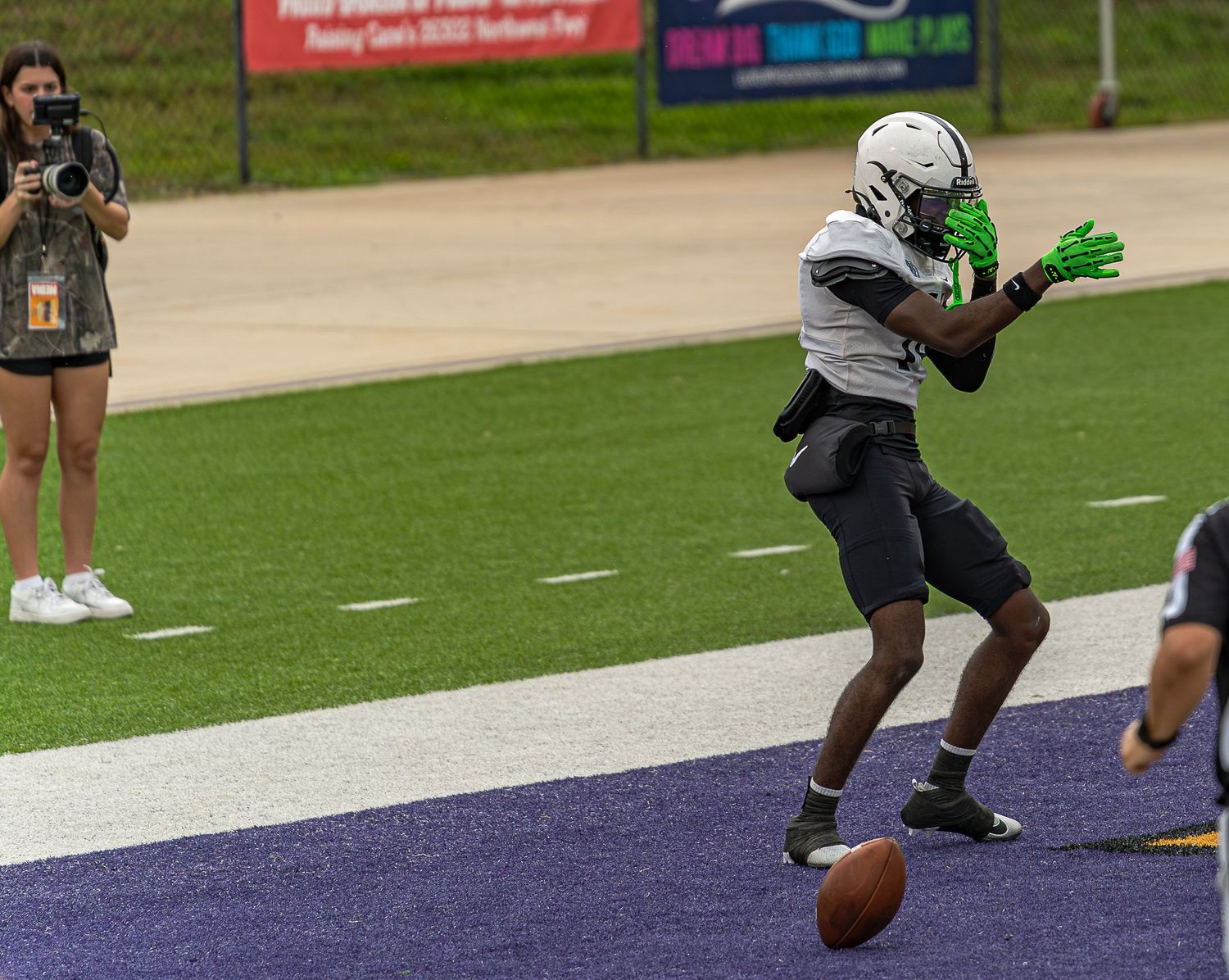 Football players run drills on a turf field during practice with purple and white endzone visible in background.