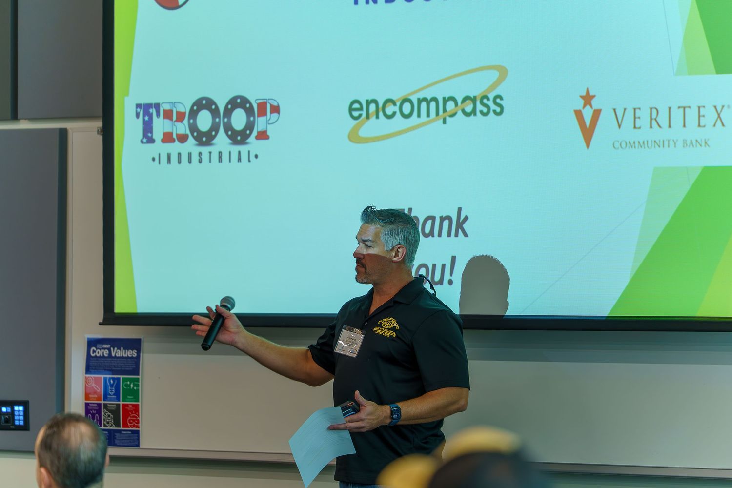 Speaker presenting in front of projection screen with corporate logos.