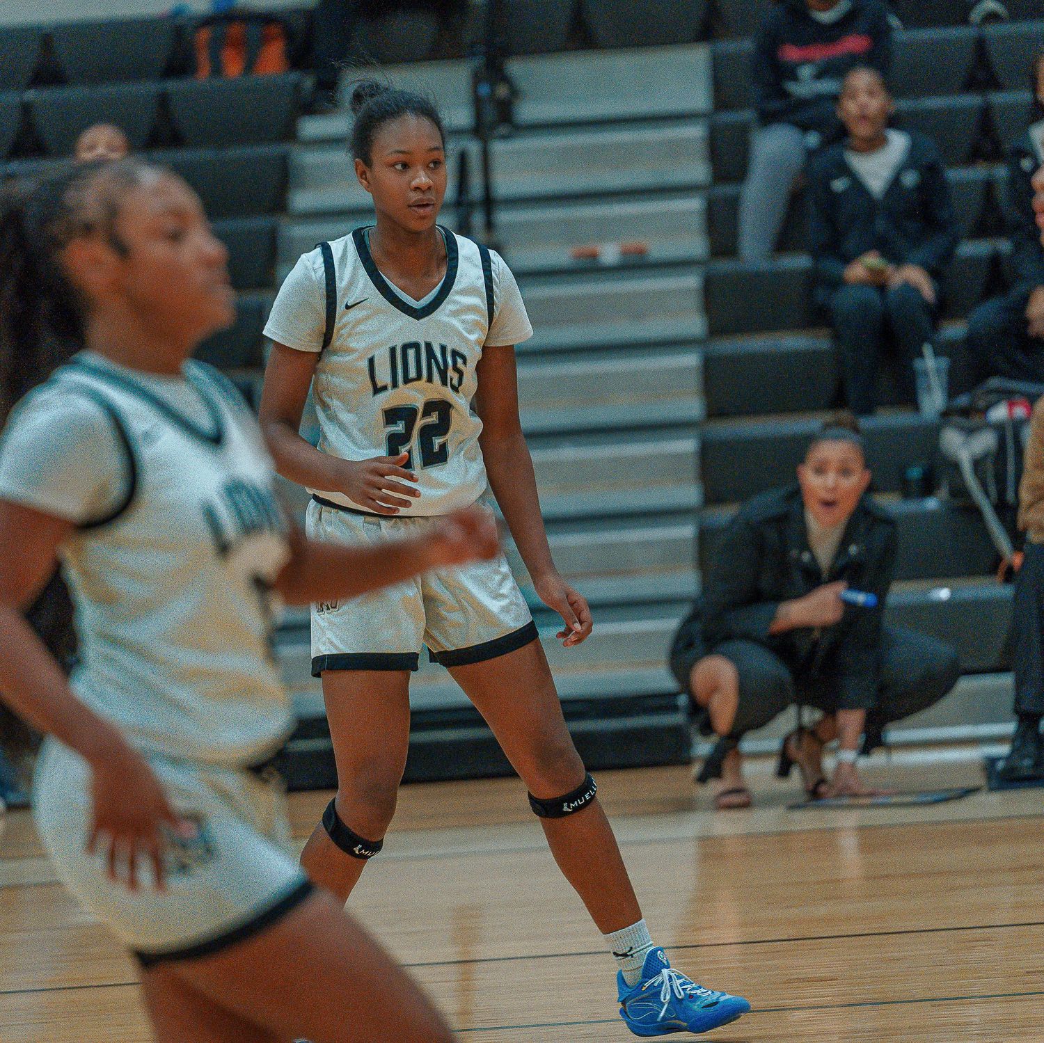 Basketball player in blue shoes crouches low in defensive position during intense game action on indoor court.