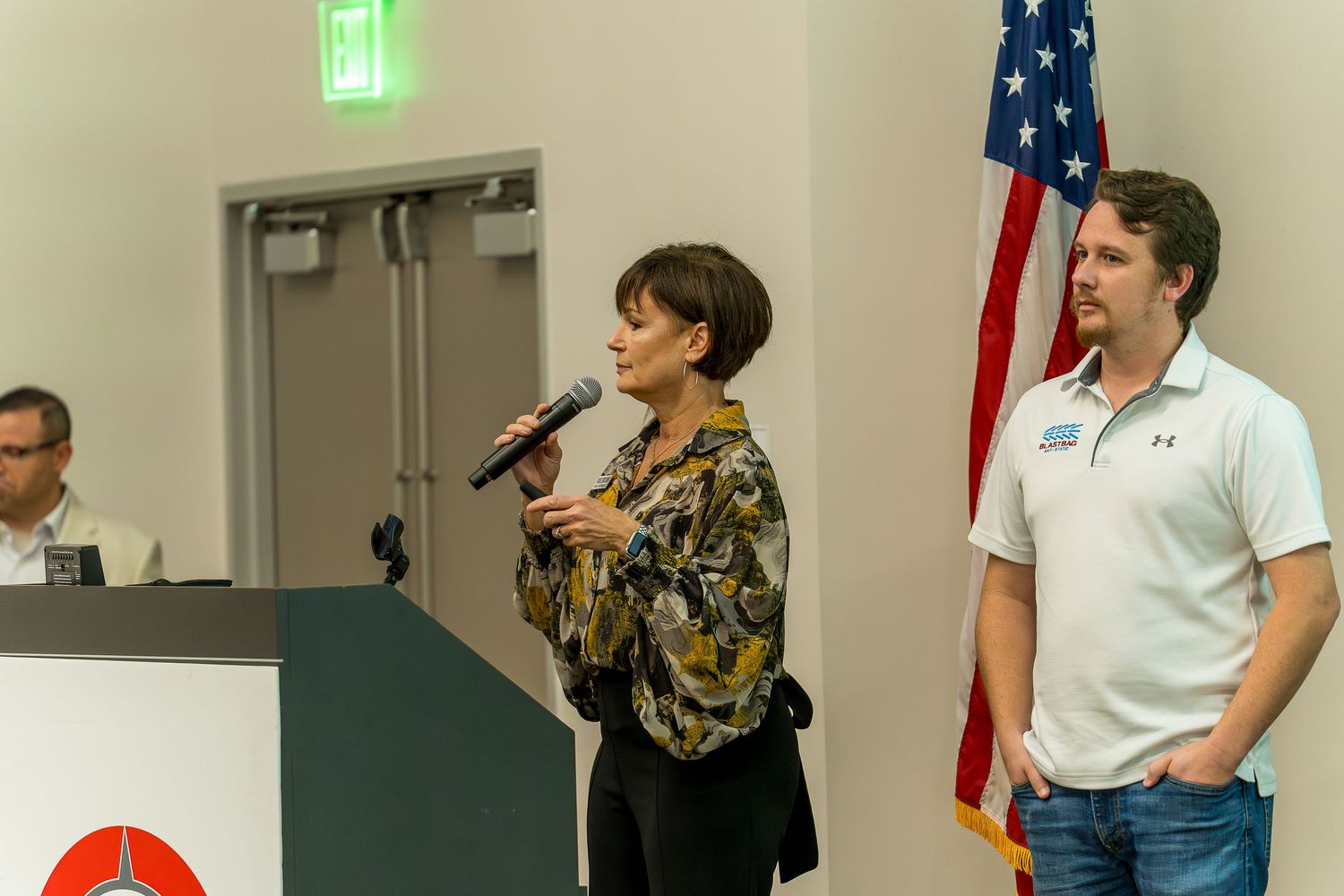 Presenters speaking at podium with American flag in background.