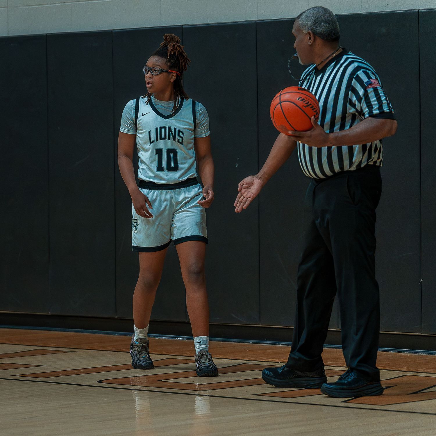 Basketball player dribbles on court during game while referee watches from sideline.