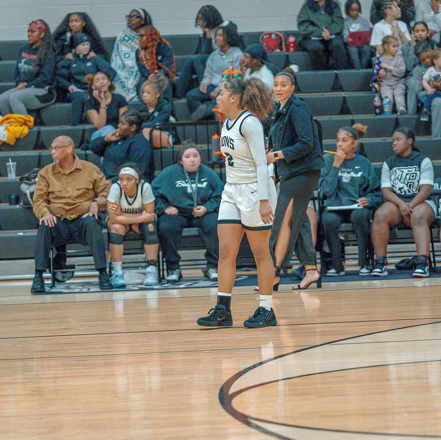 Basketball players and spectators watch from the sidelines during a game on a polished hardwood court.