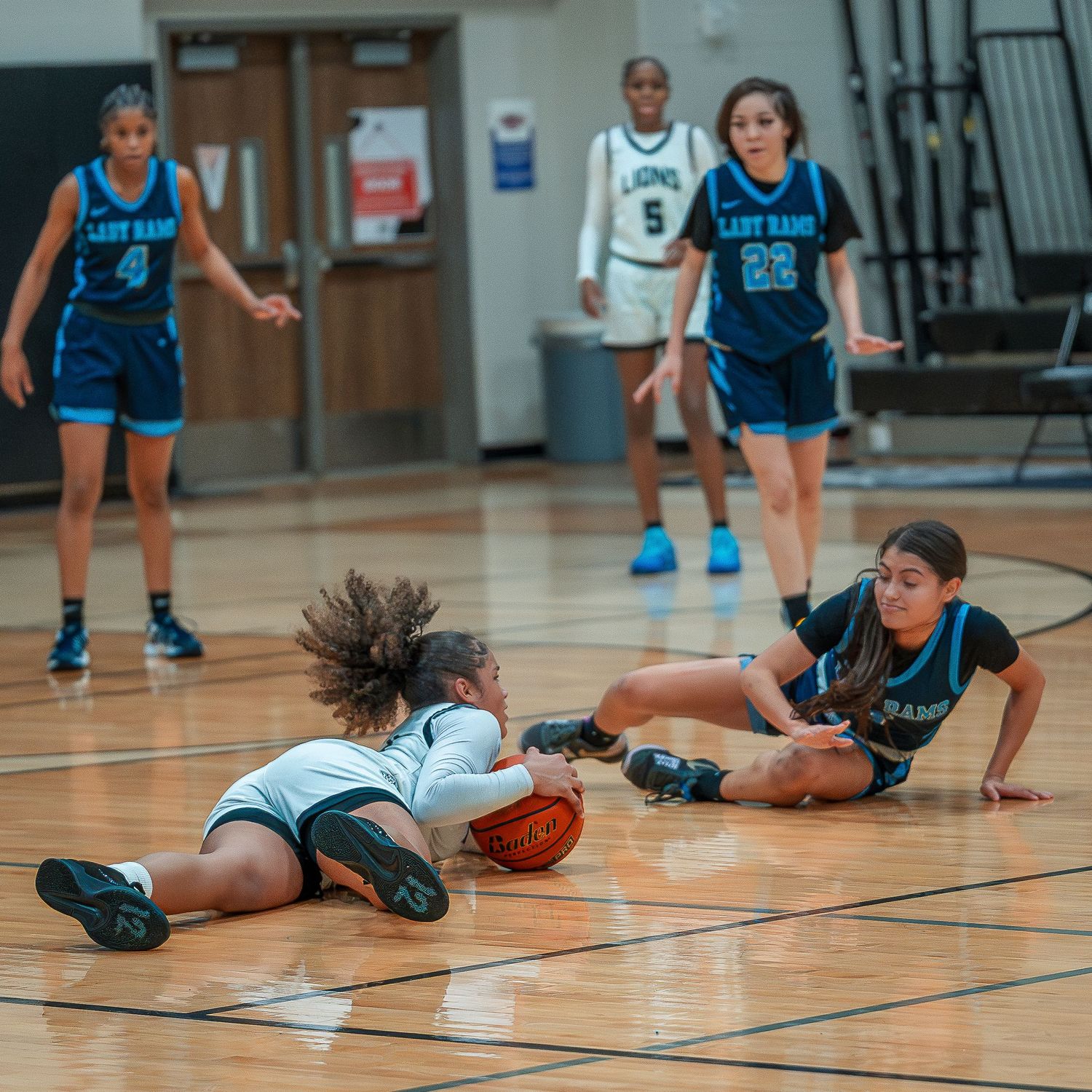 Basketball players dive and scramble for a loose ball during an intense game on a gymnasium court.