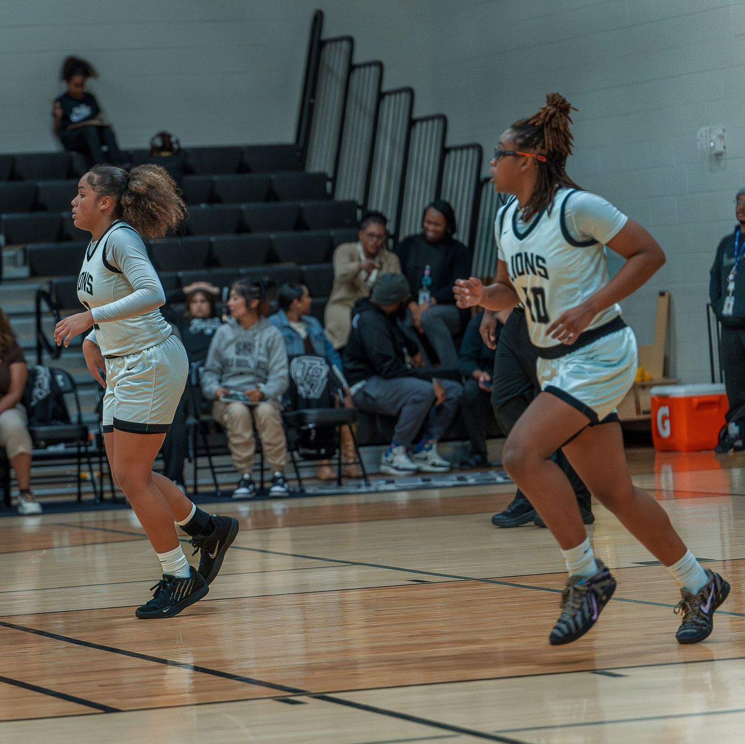 Two basketball players in white uniforms run down the court during an intense game with spectators watching from bleachers.