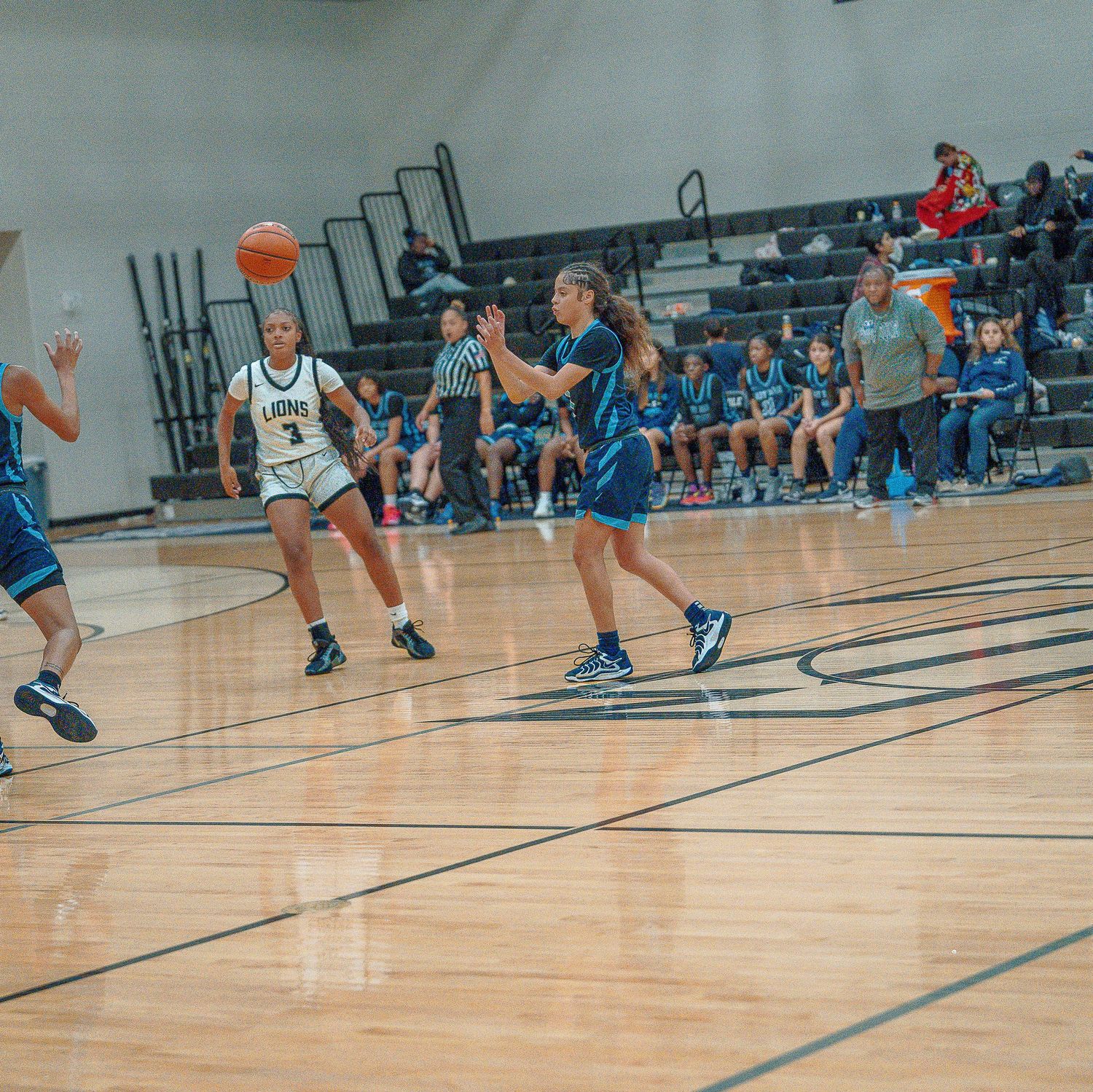 Basketball players compete in an indoor game on a wooden court with spectators watching from the sidelines.